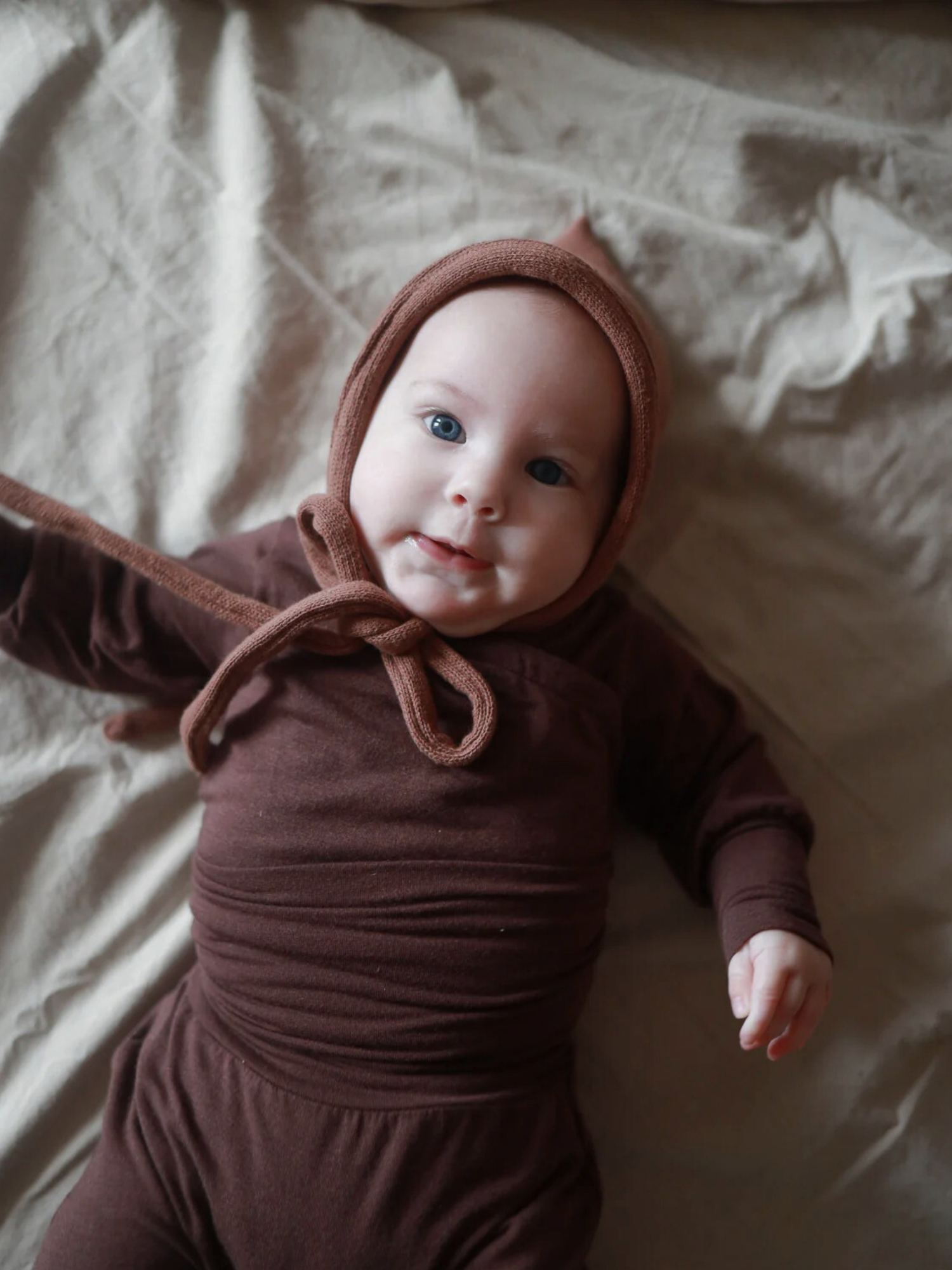 Ein Baby liegt auf einer neutralen Unterlage, gekleidet in einem Loopbody Bambusviskose - Zartbitter, inklusive passender Mütze, und schaut in die Kamera.