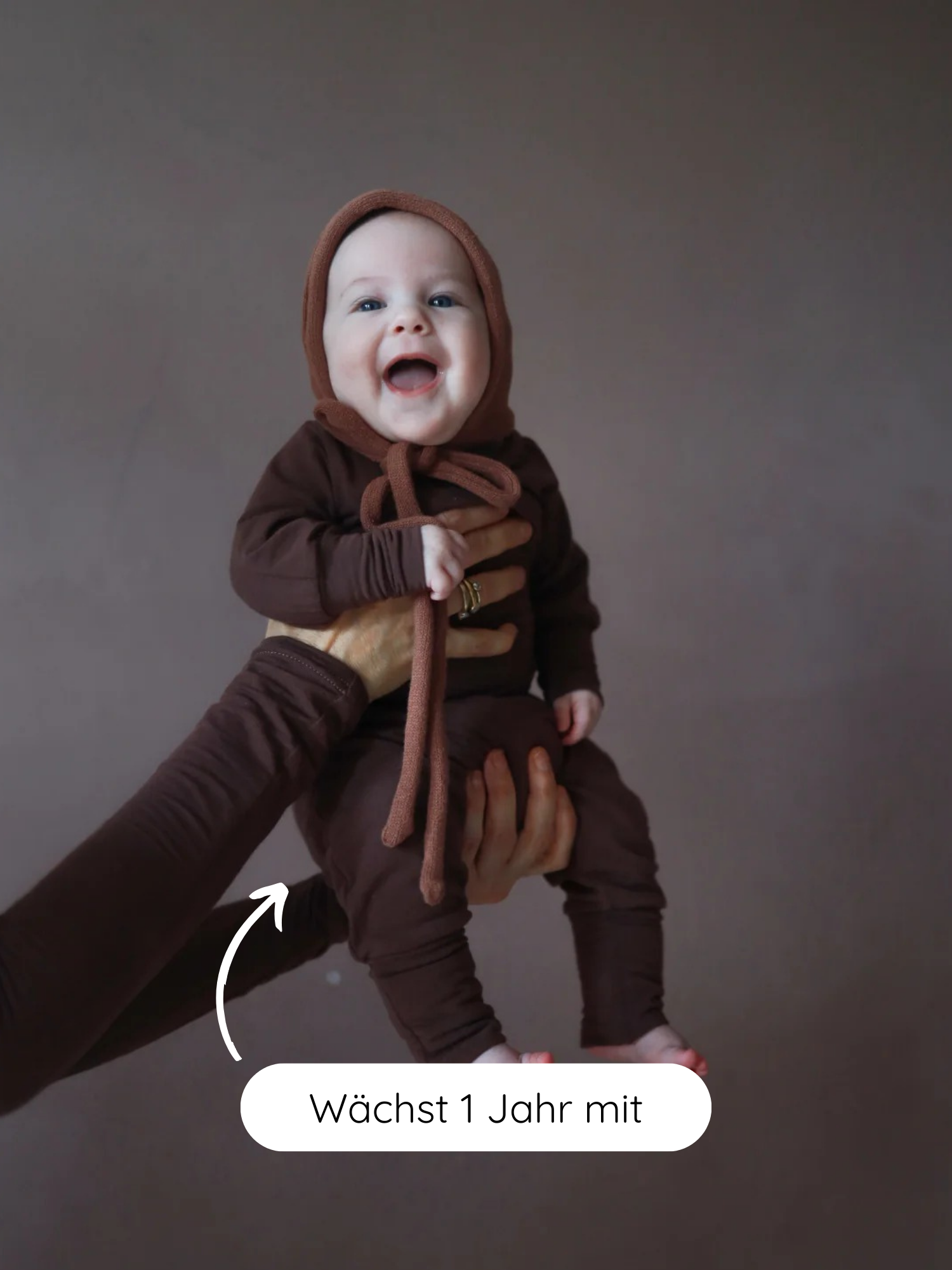Ein lächelndes Baby trägt den Loopbody Bambusviskose - Zartbitter in Schokoladenbraun, präsentiert vor einem braunen Hintergrund. Ein Pfeil und Text betonen das mitwachsende Design des bequemen, knopf- und zugfreien Babybodys.