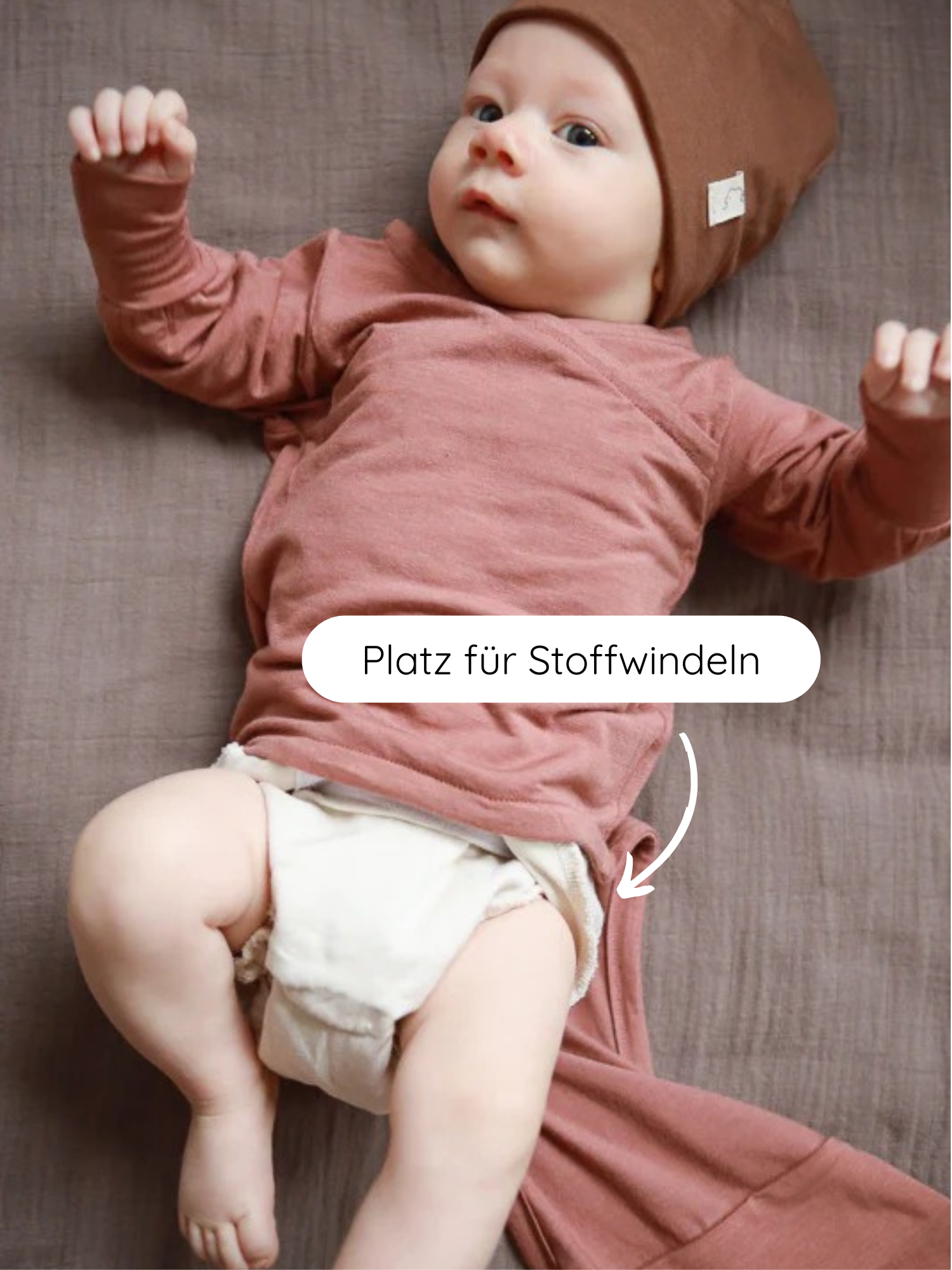 Ein Baby trägt den Loopbody Kaschmir-Mix - mitwachsender Babybody - Zimt mit passendem Hut. Eine Pfeilmarkierung zeigt auf Platz für Stoffwindeln am Body.