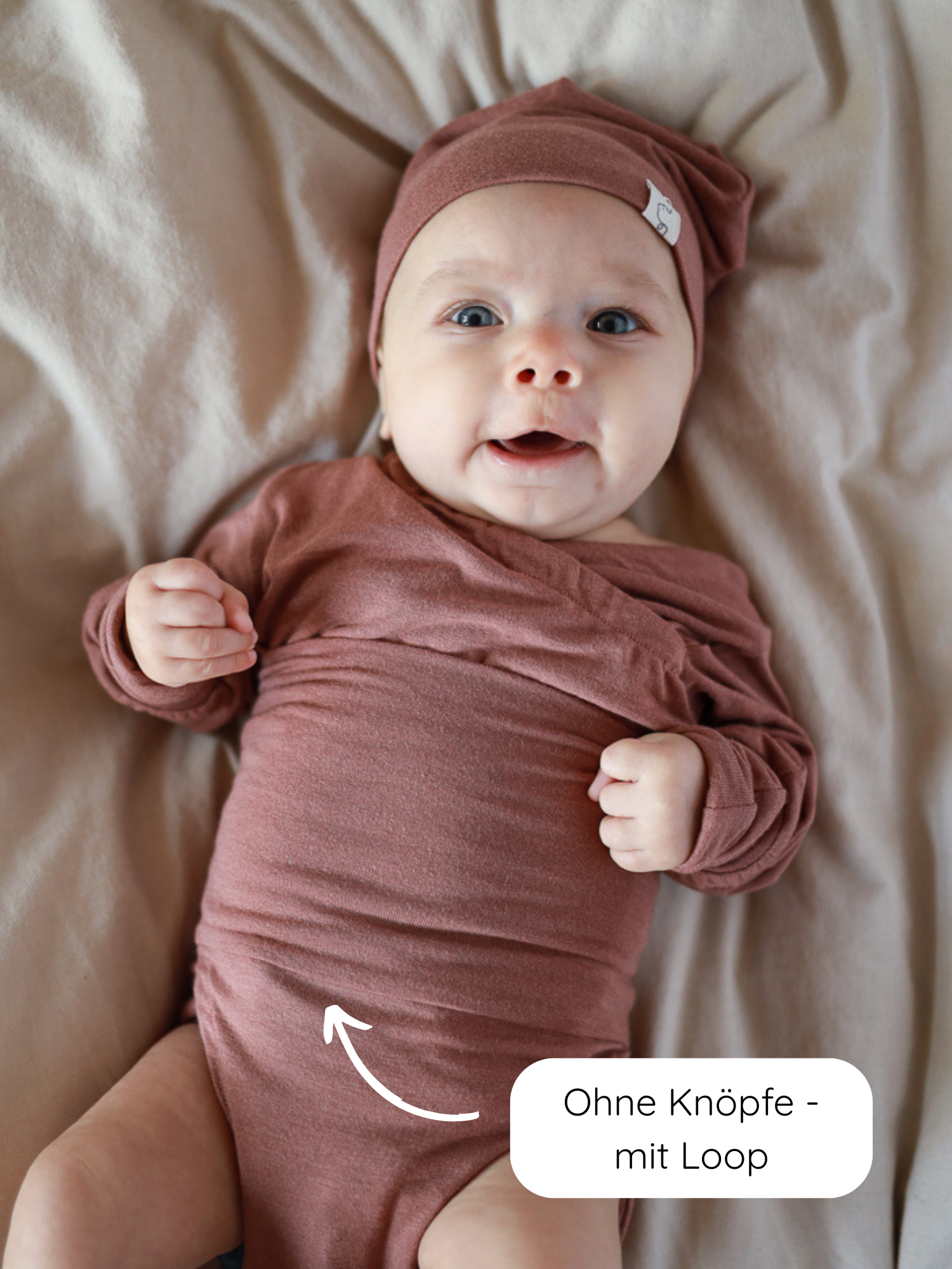 Ein Baby trägt den Loopbody Kaschmir-Mix - mitwachsender Babybody - Zimt, ohne Knöpfe, mit Loop-Verschluss, aus weichem Kaschmir-Mix, hervorgehoben durch einen Pfeil auf dem Bodysuit.