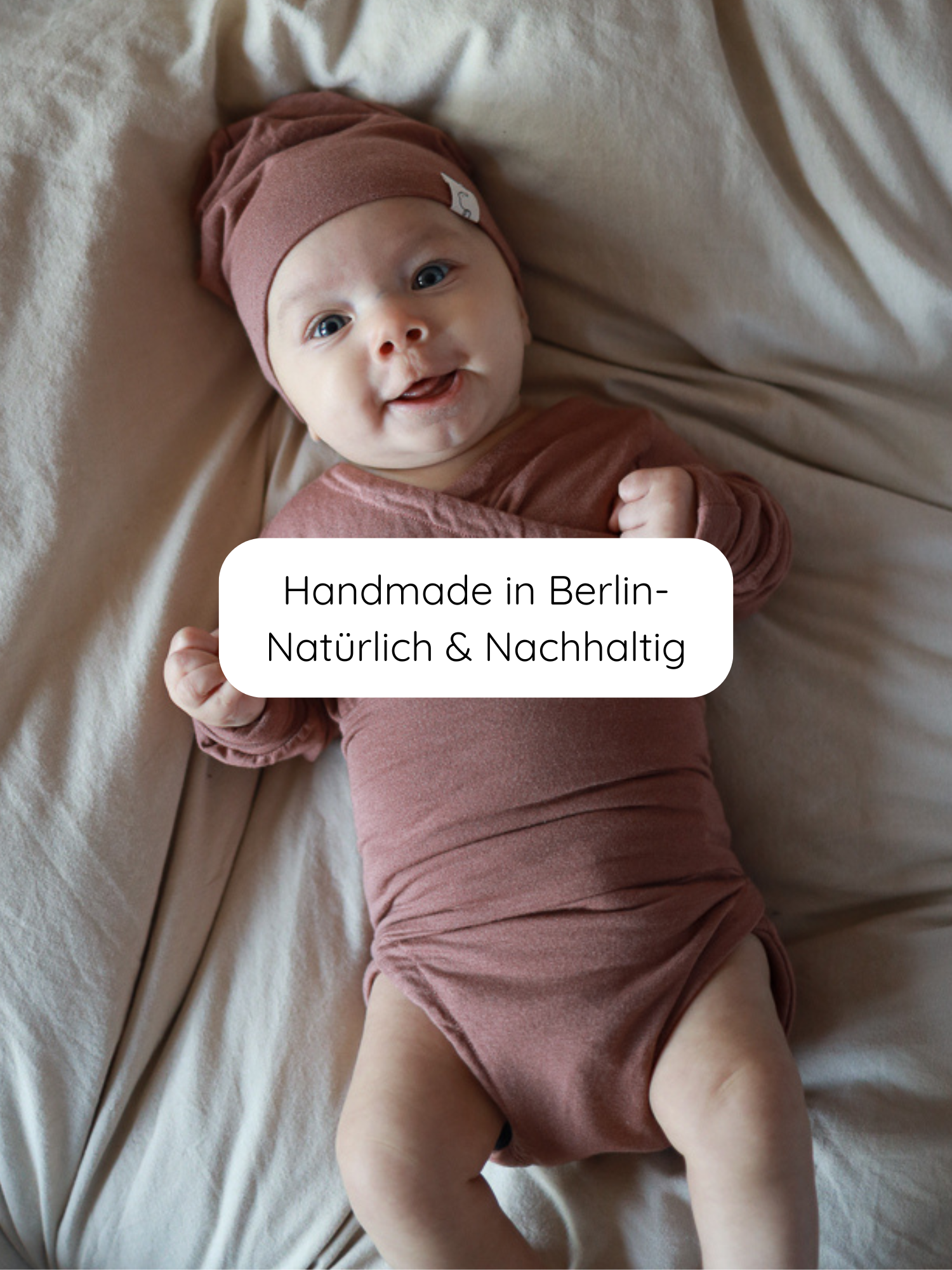 Lächelndes Baby auf hellem Laken in einem Loopbody Kaschmir-Mix - mitwachsender Babybody - Zimt, mit passender Mütze, handgefertigt in Berlin, natürlich und nachhaltig.