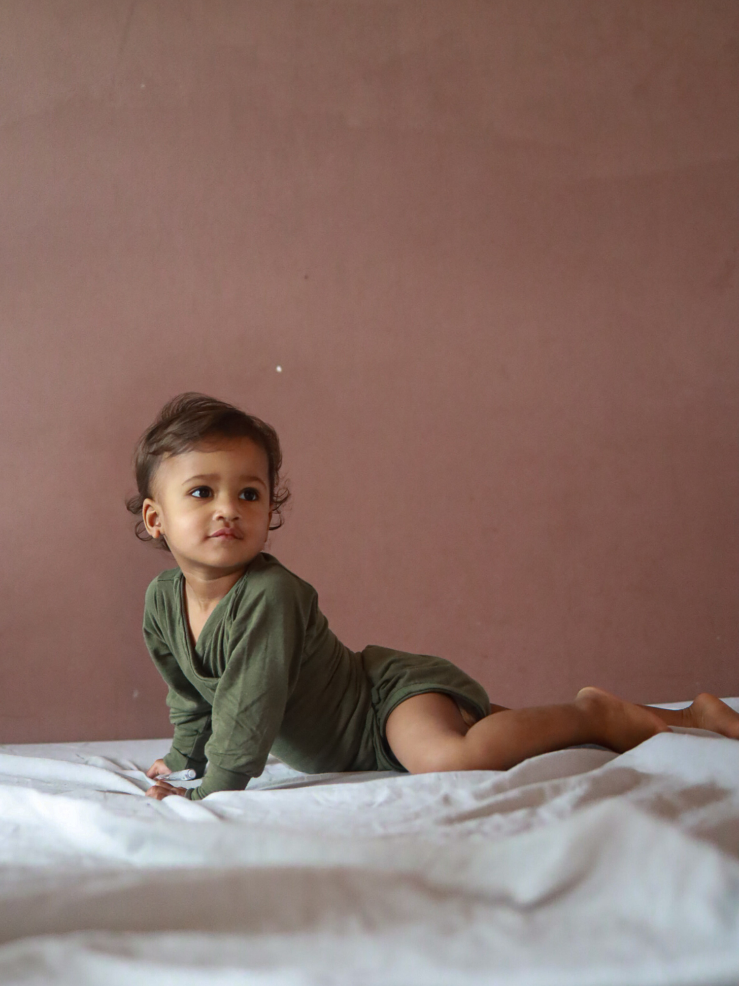 Kleinkind in einem Loopbody Kaschmir-Mix - Avocado, langärmelig und ohne Knöpfe, auf einem Bett krabbelnd. Der Body passt sich dem Baby an und bietet Komfort und Wärme.