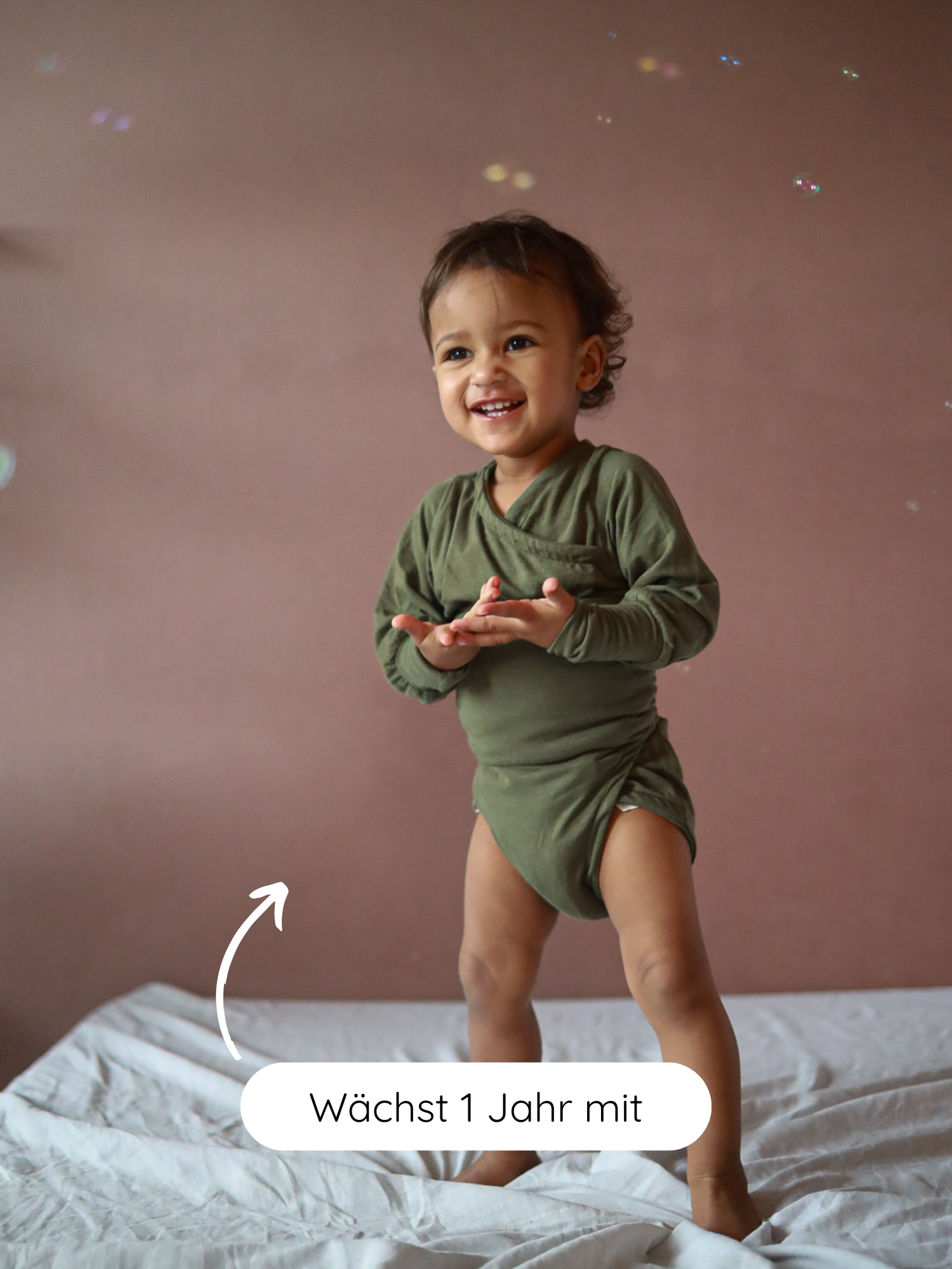 Kleinkind in einem Loopbody Kaschmir-Mix - Avocado, spielt fröhlich mit Seifenblasen. Der langärmelige Body wächst ein Jahr mit dem Baby und bietet Komfort und Wärme.