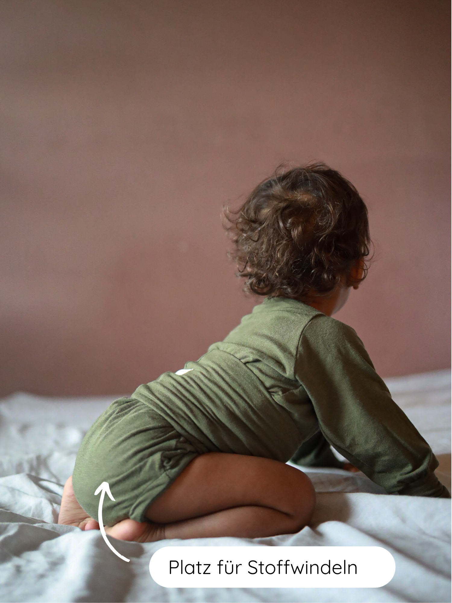 Kleinkind in einem Loopbody Kaschmir-Mix - Avocado aus Kaschmir-Mix, das auf einem Bett krabbelt, mit Pfeil zur großzügigen Sitzfläche für Stoffwindeln.