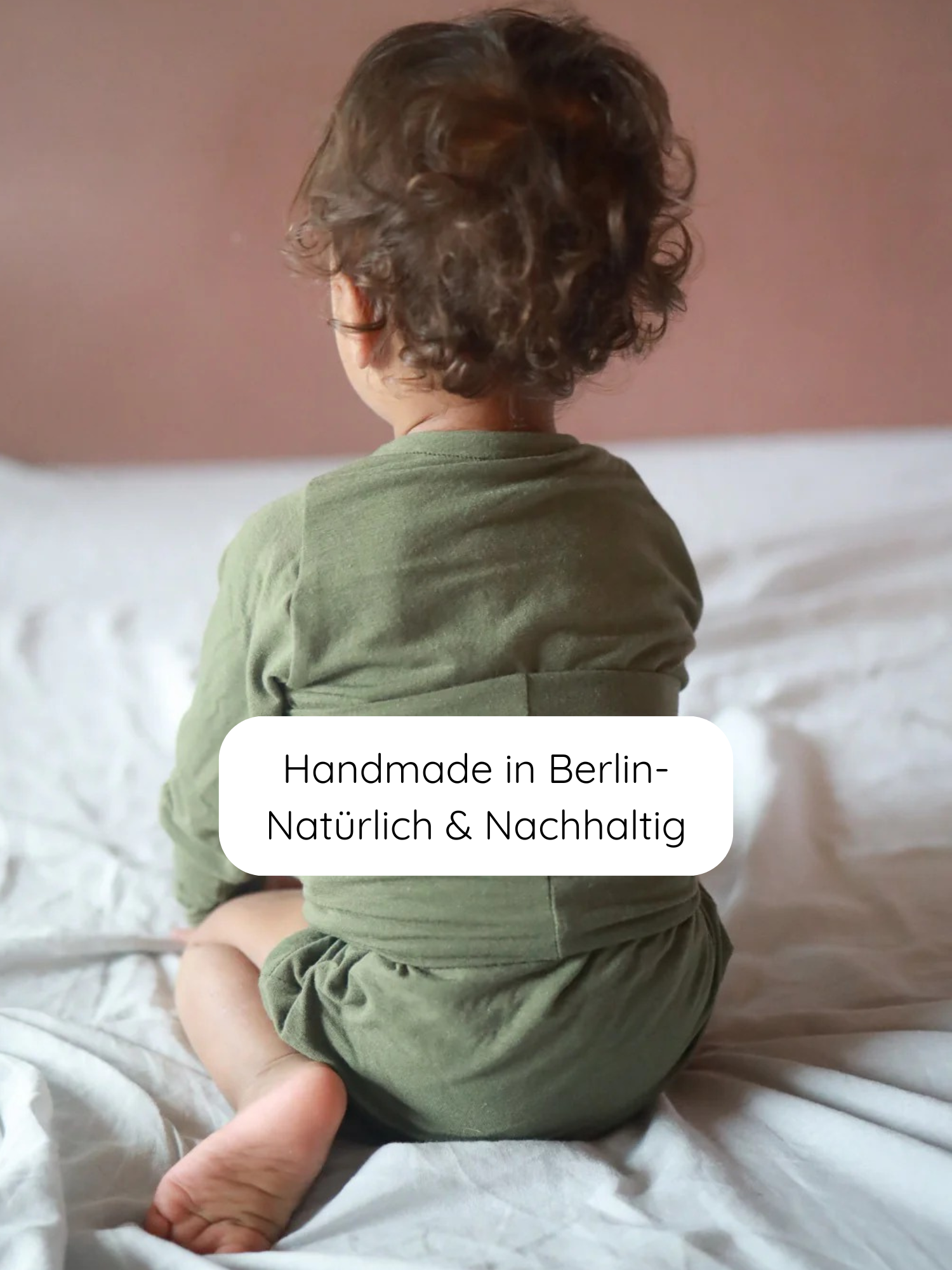 Kleinkind von hinten auf heller Decke, trägt einen Loopbody Kaschmir-Mix - Avocado. Handgefertigt in Berlin, nachhaltig und bequem, ohne Knöpfe, mitwachsendes Design für maximalen Tragekomfort.