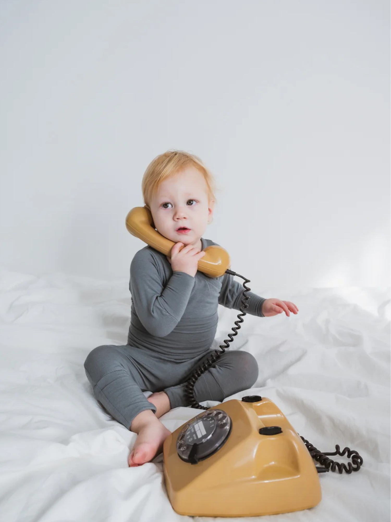 Kleinkind auf weißem Bett trägt Babyknicker Kaschmir-Mix - Taube mit Klappbündchen, hält Vintage-Telefonhörer ans Ohr, sitzt vor gelbem Retro-Telefon. Komfortabel und weich, ideal für Neugeborene.