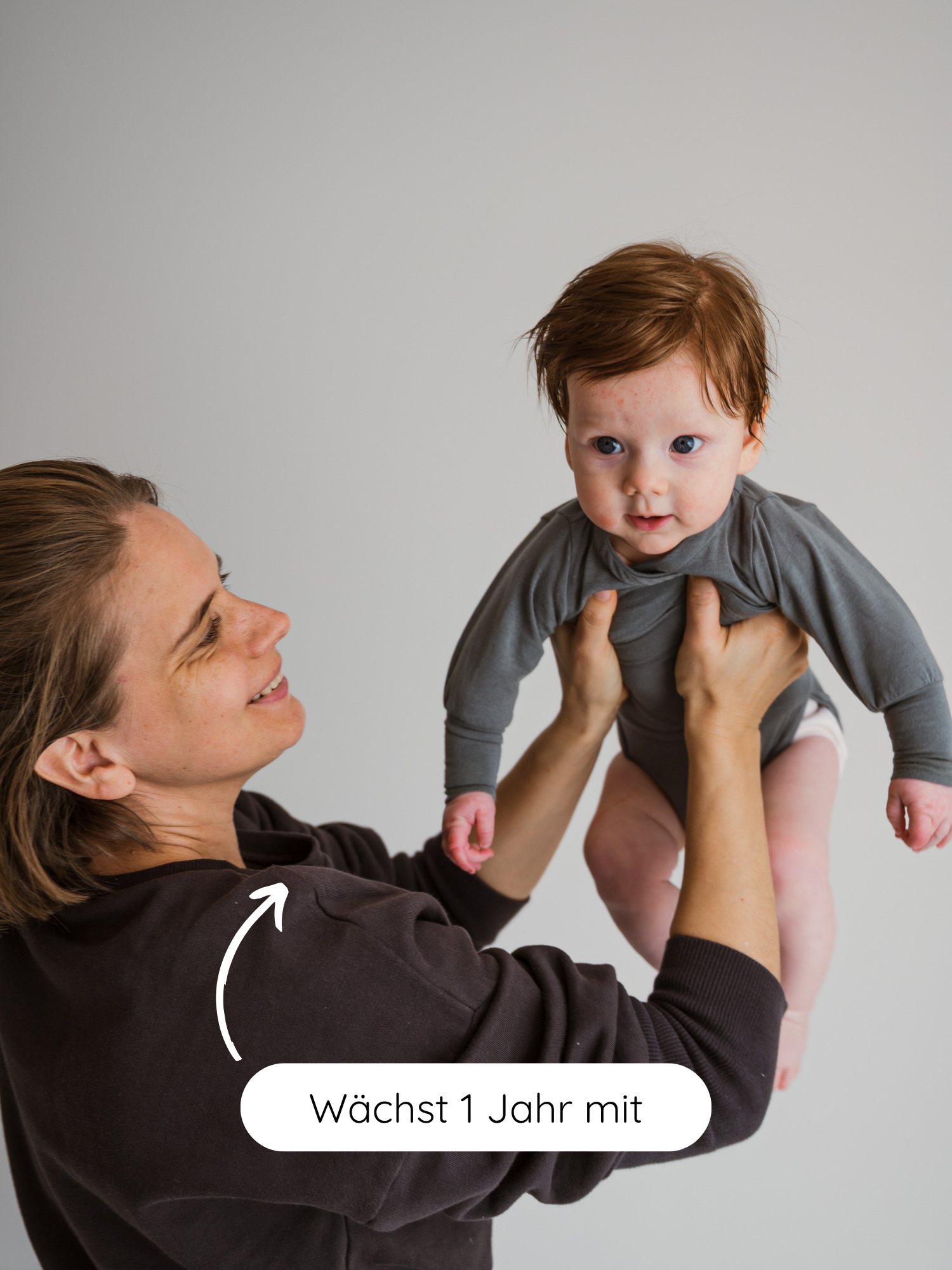 Erwachsene hält Baby in grauem Loopbody Kaschmir-Mix - Taube, der ein Jahr mitwächst, hoch. Der Body bietet Komfort, Wärme und ist ideal für empfindliche Babyhaut.