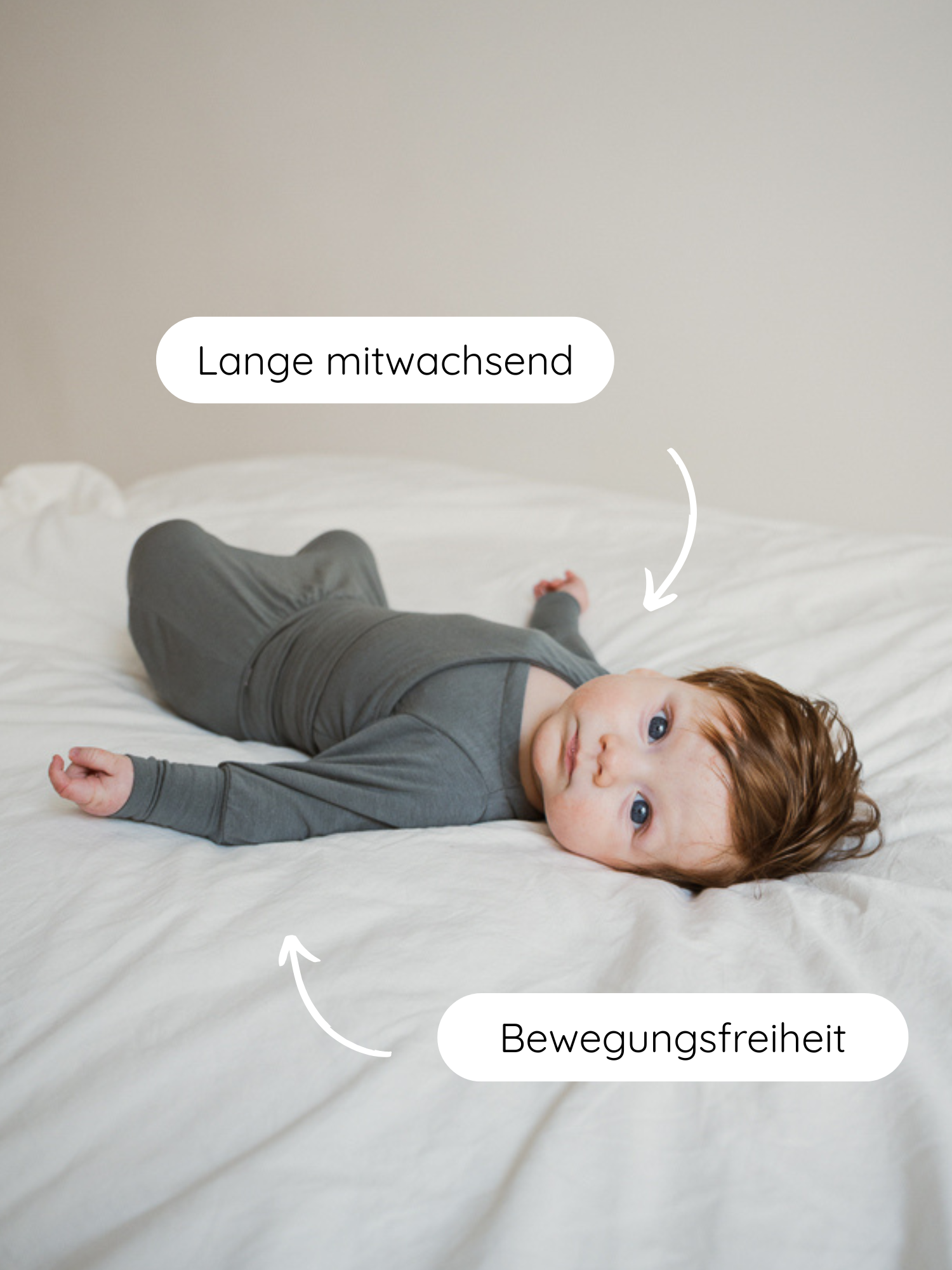 Baby liegt auf weißem Bett, trägt einen anliegenden, schiefergrauen Babyknicker Kaschmir-Mix - Taube. Pfeile zeigen auf Lange mitwachsend am Torso und Bewegungsfreiheit an Armen und Beinen.