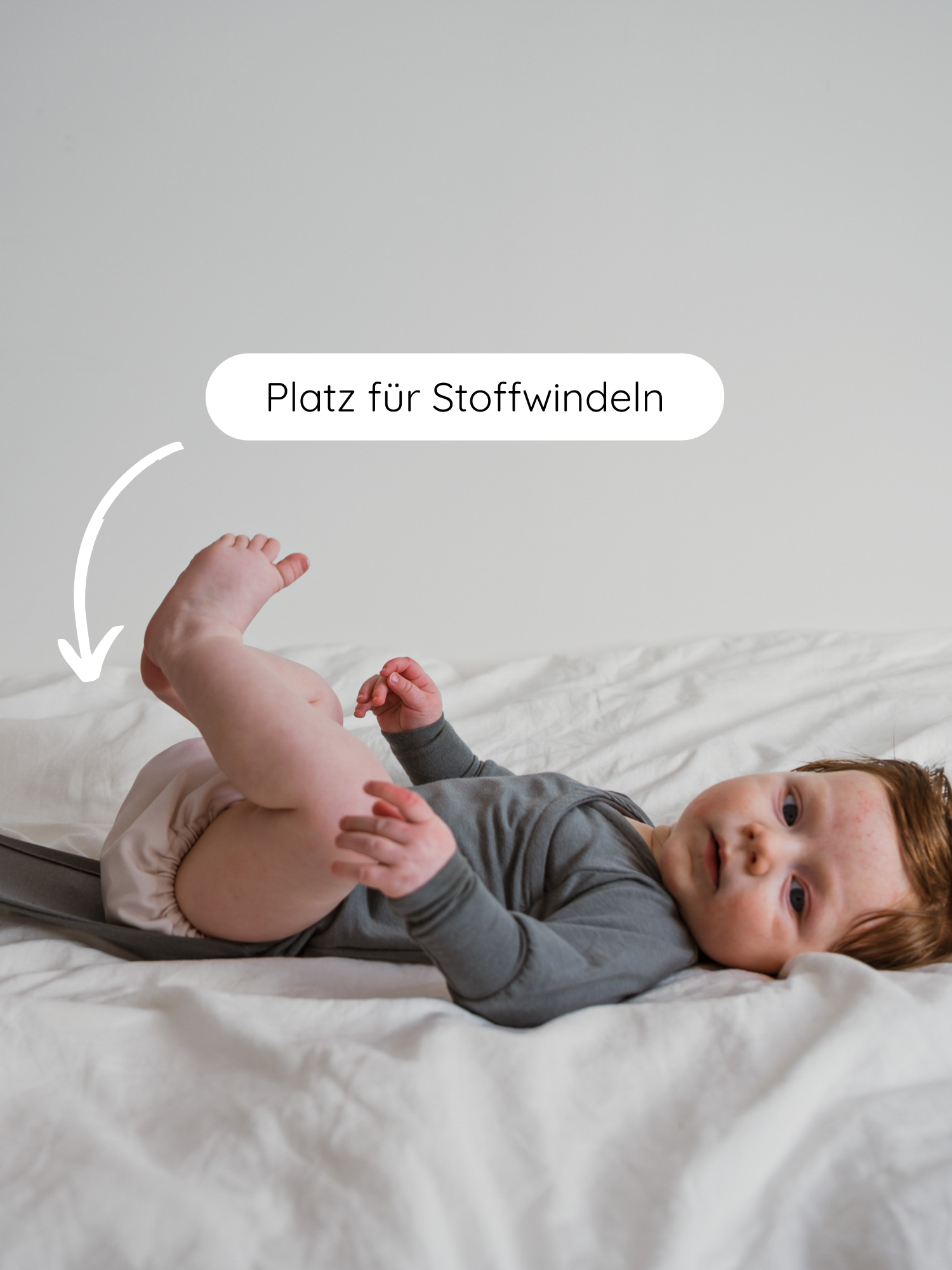 Säugling auf weißem Bett in grauem Loopbody Kaschmir-Mix - Taube, trägt hellen Stoffwindel. Weißer Pfeil und Text Platz für Stoffwindeln zeigen die Windelanpassungsfähigkeit.