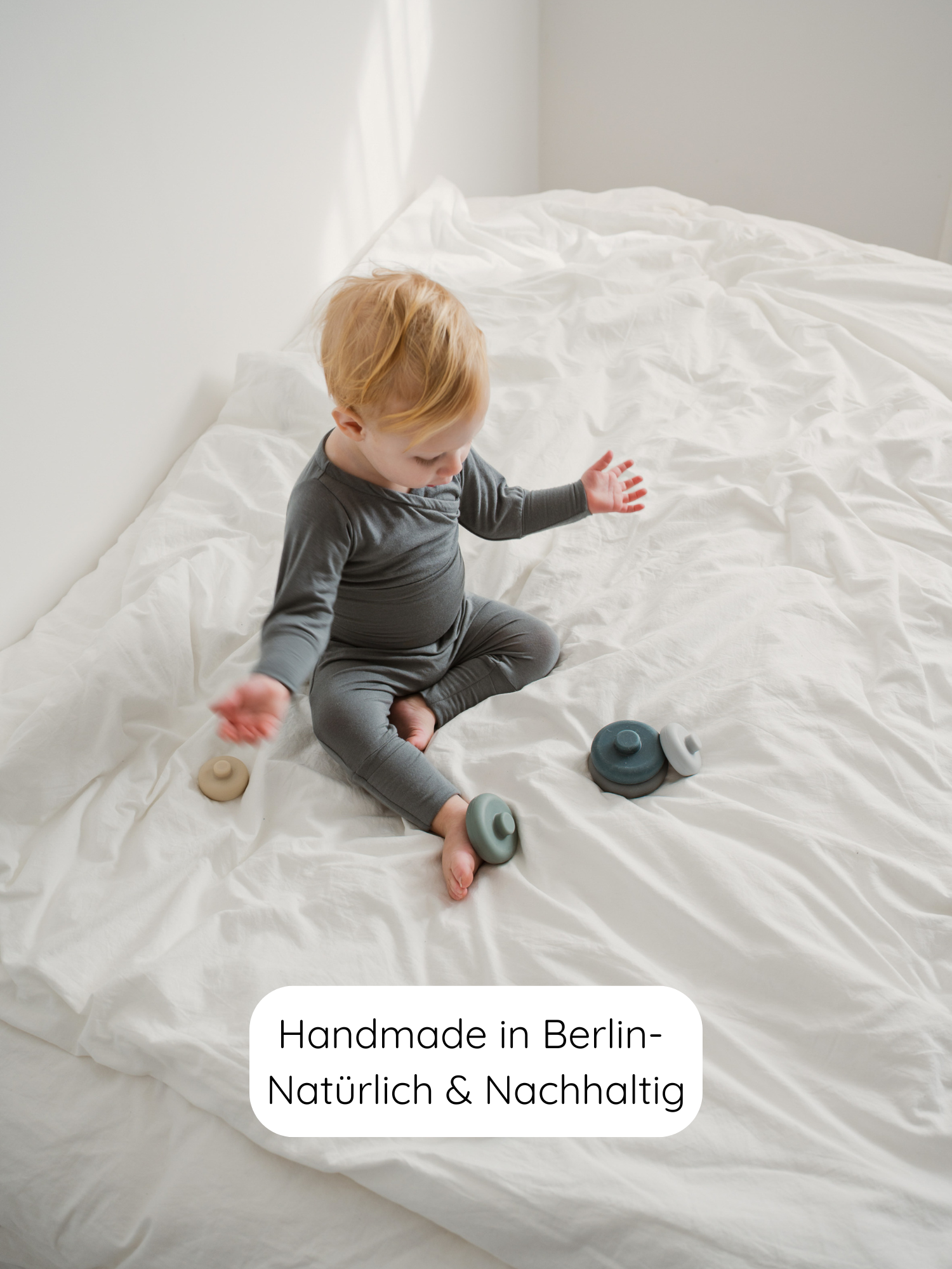 Kleinkind in grauem Loungewear auf weißem Bett mit Babyknicker Kaschmir-Mix - Taube, umgeben von gestapelten Spielzeugen. Label: Handmade in Berlin – Natürlich & Nachhaltig.