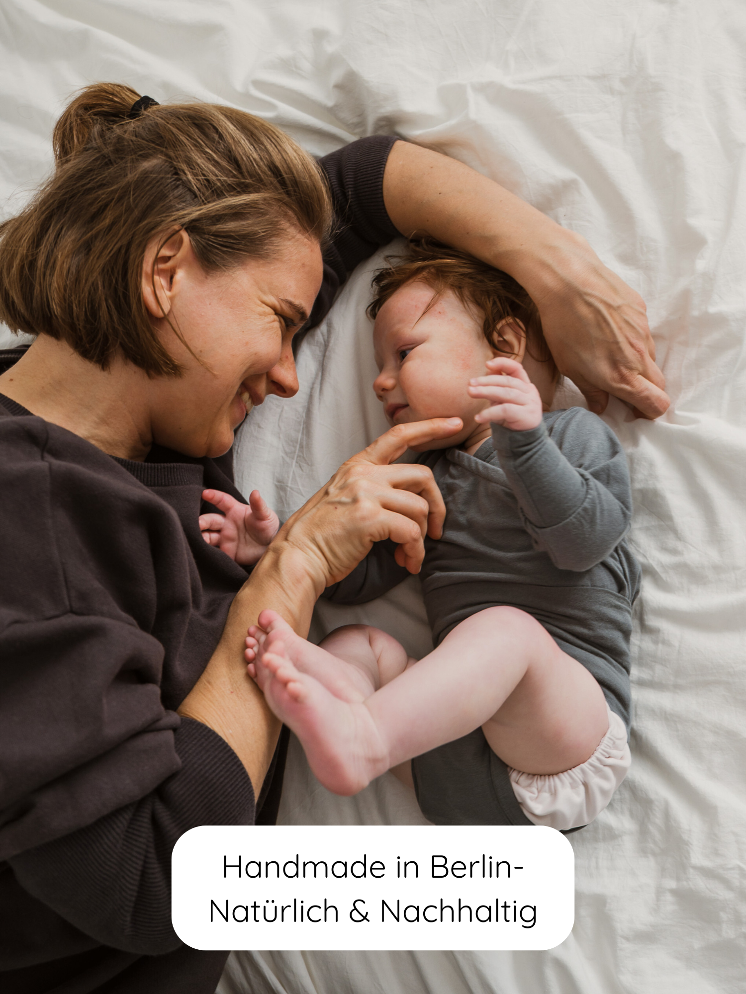 Mutter lächelt Baby im grauen Loopbody Kaschmir-Mix an, der weich und bequem ist, ohne Knöpfe. „Handmade in Berlin – Natürlich & Nachhaltig.“ steht unten.