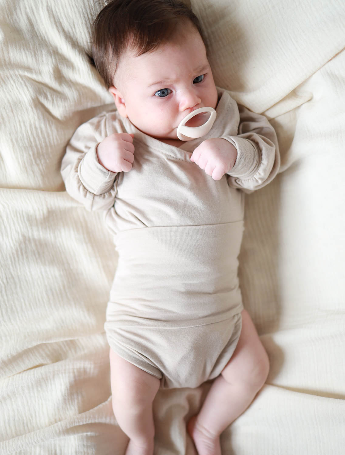 Baby liegend auf einer Decke trägt den Loopbody Kaschmir-Mix – Sand, ohne Knöpfe, mit Loop-Design, das den Bauch umgibt, aus einem weichen Kaschmir-Modal-Mix. Erstausstattung Baby und Baby Geschenk zur Geburt.