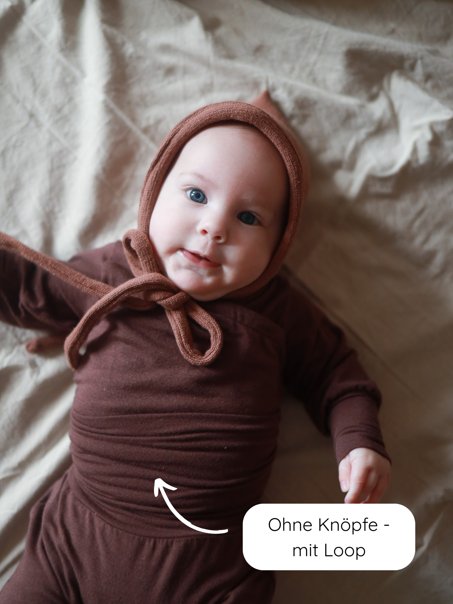 Loopbody Kaschmir-Mix - Malve: Baby in braunem Strickoutfit mit passender Mütze auf cremefarbenem Laken. Ein Pfeil zeigt auf den knopf- und reißverschlussfreien Loop-Body für einfaches An- und Ausziehen.