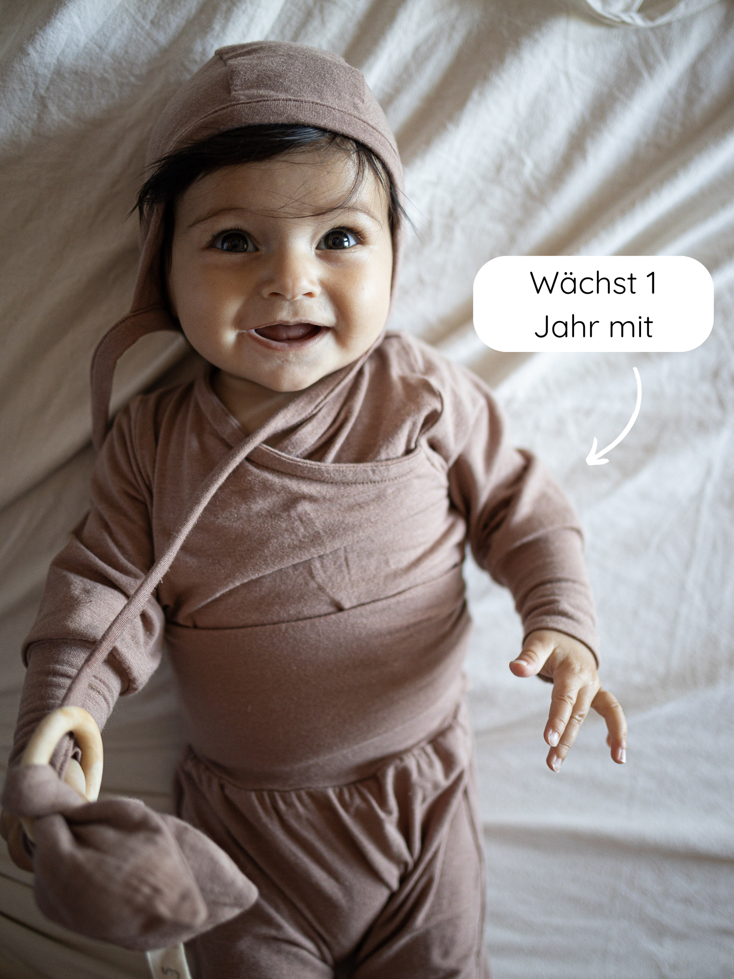 Ein lächelndes Baby trägt den Loopstrampler Kaschmir-Mix - Puder, eine taupefarbene, knopflose Baumwollbekleidung, mit passender Mütze und hält ein Holzspielzeug mit Stoffohren.