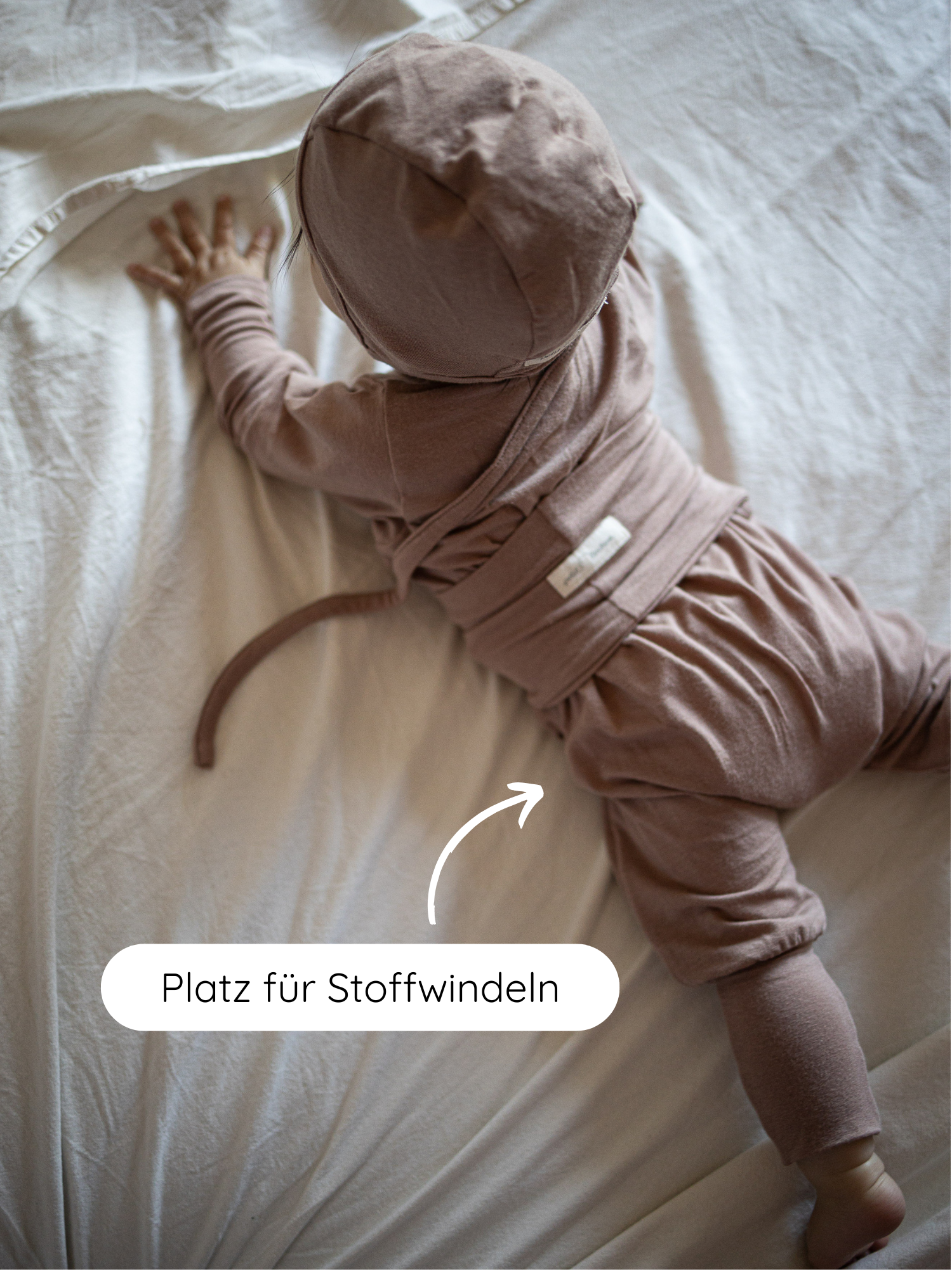 Kleinkind in Loopstrampler Kaschmir-Mix - Puder, einem weichen, knopflosem Outfit mit Platz für Stoffwindeln. Bonnet, Wickeloberteil, lockere Hose und Beinstulpen bieten Komfort und Wärme.