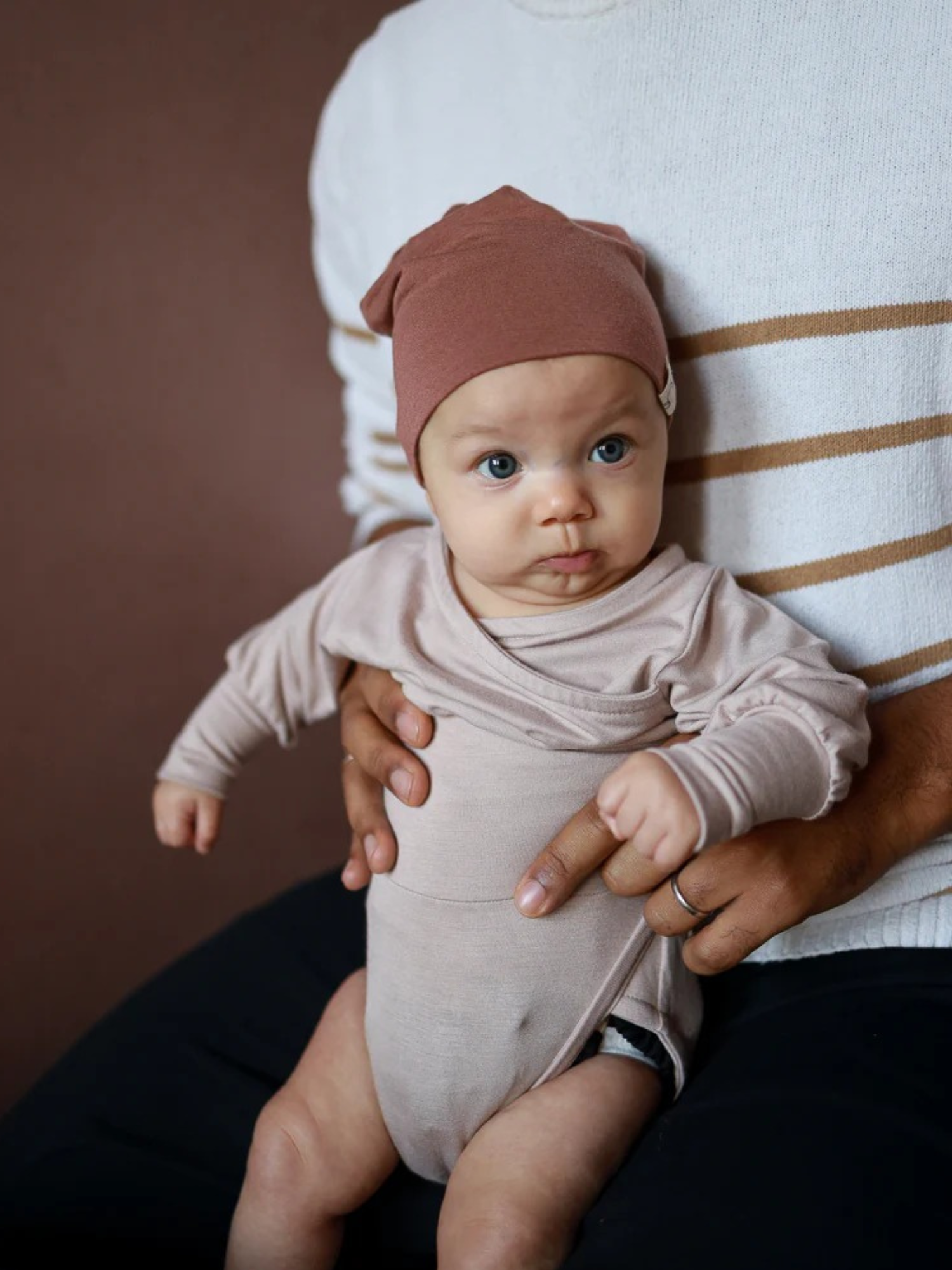Ein Baby im Loopbody Bambusviskose - Cremepuder sitzt auf dem Schoß eines Erwachsenen, der es sanft hält. Der Loopbody bietet Komfort und Schutz durch sein weiches, mitwachsendes Design.