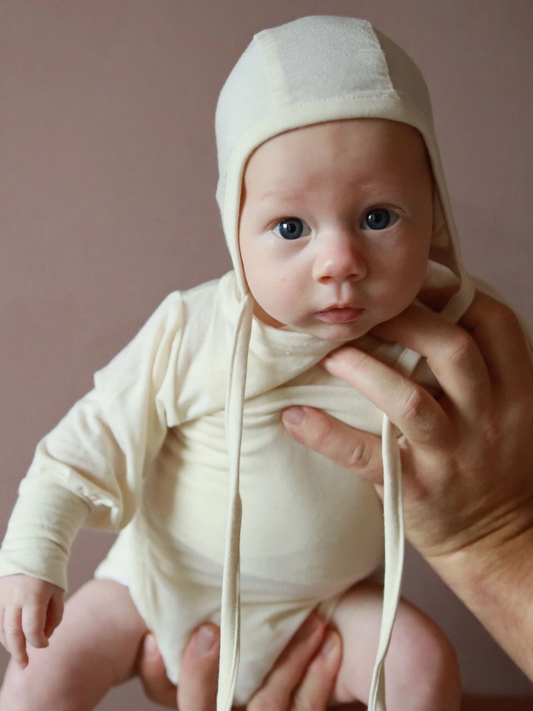 Kleines Baby in weich-cremefarbenem Loopbody Bambusviskose - Natur und passender Haube, gehalten von Erwachsenen. Das Design ist bequem, wärmend und mitwachsend, ideal für empfindliche Babyhaut.