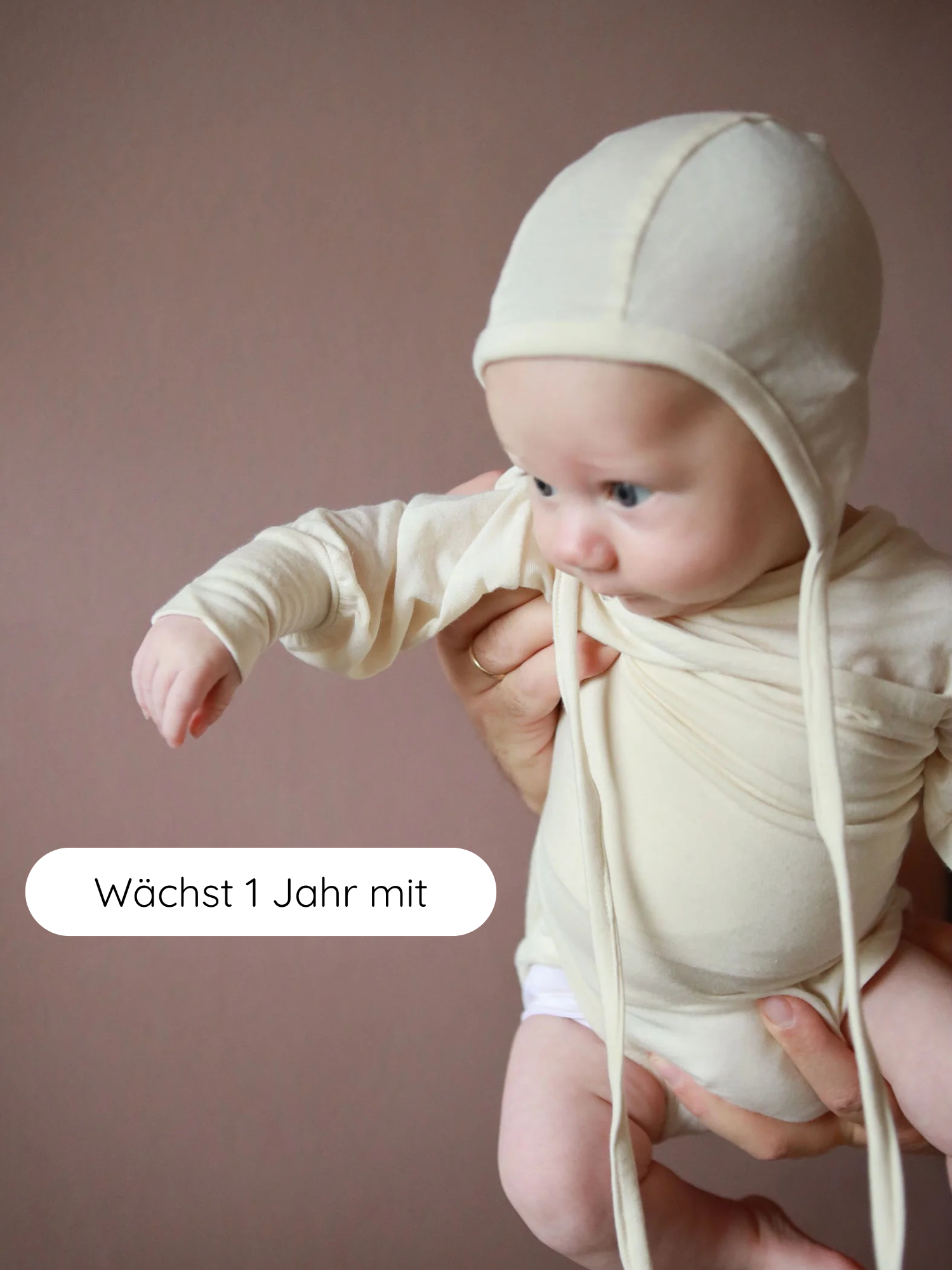 Kleines Baby im einteiligen Loopbody Bambusviskose - Natur mit passender Haube, gehalten von Erwachsenen. Text „Wächst 1 Jahr mit“ betont das mitwachsende Design des bequemen, atmungsaktiven Babybodys.