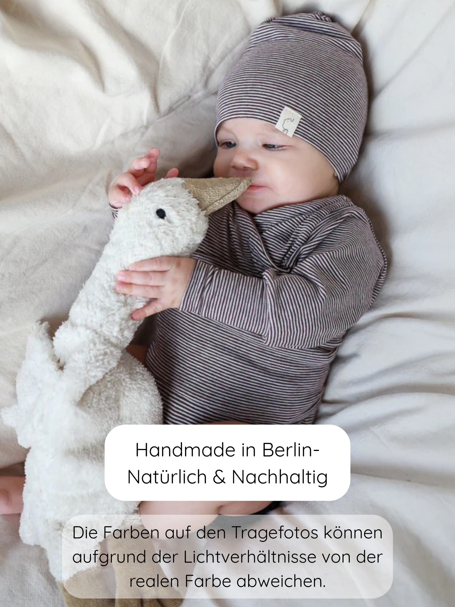 Baby in einem Loopbody Bio-Baumwolle-Ringel Schoko mit passender Mütze, spielt mit einem Plüschgans-Spielzeug. Über dem weichen Untergrund sind deutsche Beschreibungen zur Herstellung und Farbabweichungen eingeblendet.