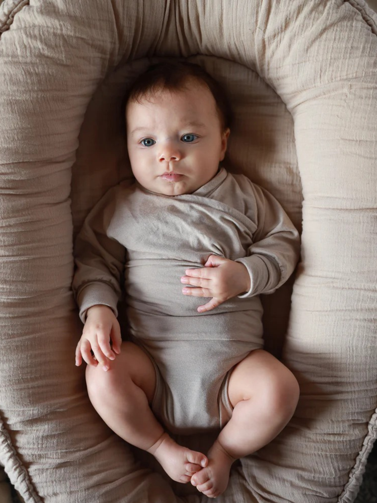 Baby liegt in einem weichen, beigen gepolsterten Nest, gekleidet in den Loopbody Kaschmir-Mix - Sand, einem langärmeligen, wickelstil Body, der eine gemütliche und geschützte Umgebung bietet.