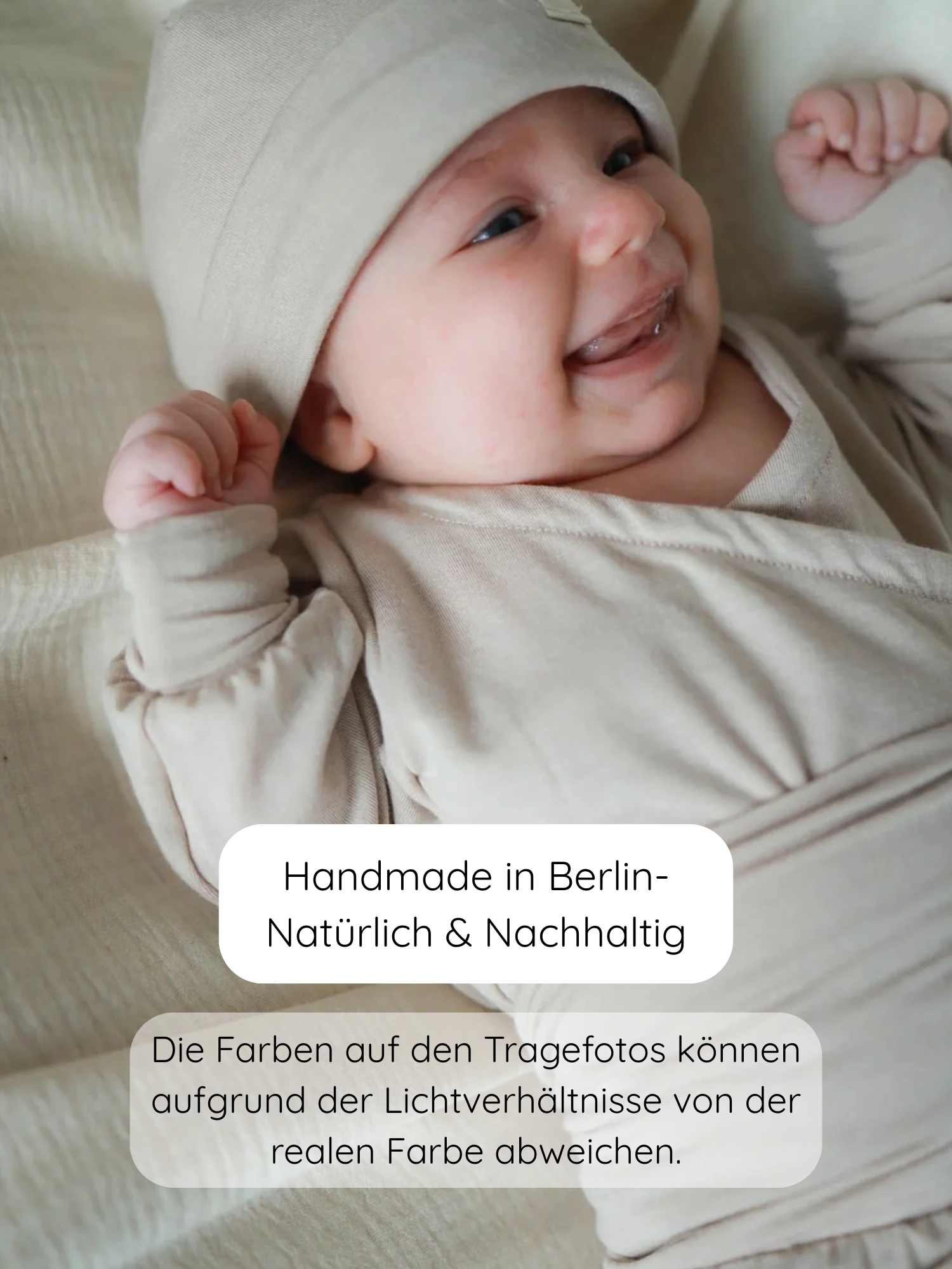 Ein lächelndes Baby trägt den Loopbody Kaschmir-Mix in Sand, ergänzt durch eine passende Mütze, auf neutralem Stoff liegend. Handgemacht in Berlin; Farben können leicht abweichen.