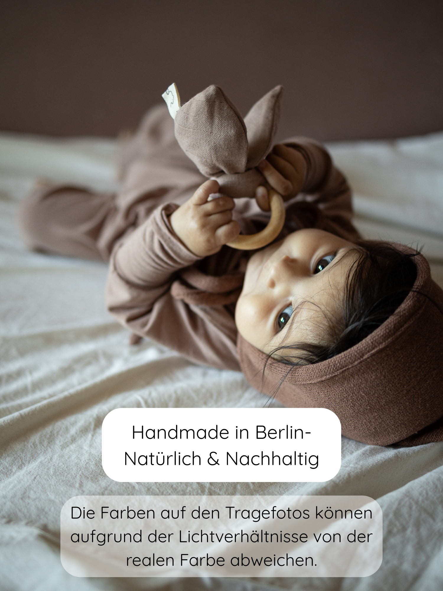 Ein Baby liegt auf einem weichen Untergrund, trägt den Loopstrampler Kaschmir-Mix - Puder mit passendem braunem Outfit und Bonnet, hält einen hölzernen Beißring mit Stoffohren, nachhaltig handgefertigt in Berlin.