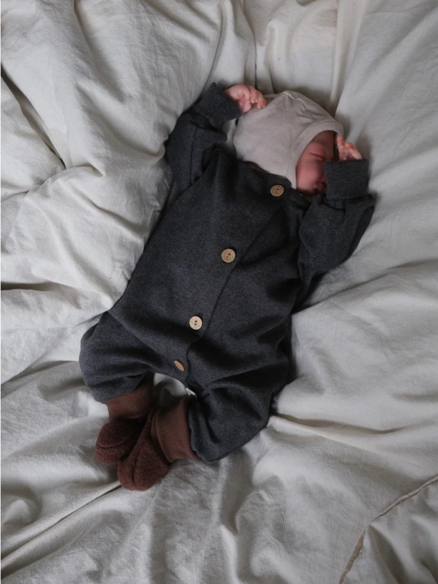 Neugeborenes in einem Mini Overall Feinstrick - Ruß Meliert aus Baumwolle-Elasthan, schlafend auf einer weißen Decke. Der Overall ist bequem, mit Knöpfen vorne, ideal zum Schlafen und Spielen.