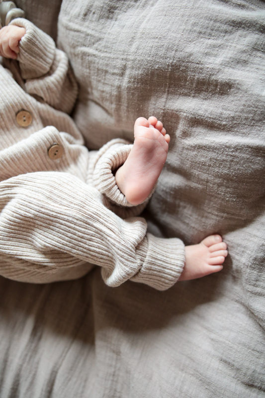 Baby liegt auf dem Bett. Trägt Strampler mit nackten Füßchen. Baby Erstausstattung und Baby Geschenk von Petit Cochon.