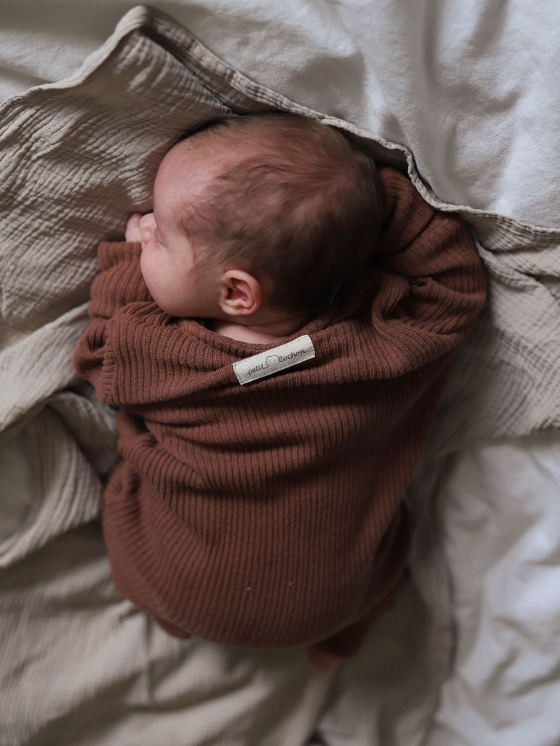 Ein Baby schläft auf einer Decke, eingewickelt in den weichen Mini Overall Rippstrick aus recycelter Baumwolle, der Komfort und Wärme bietet. Der nachhaltige Strampler für Babys.