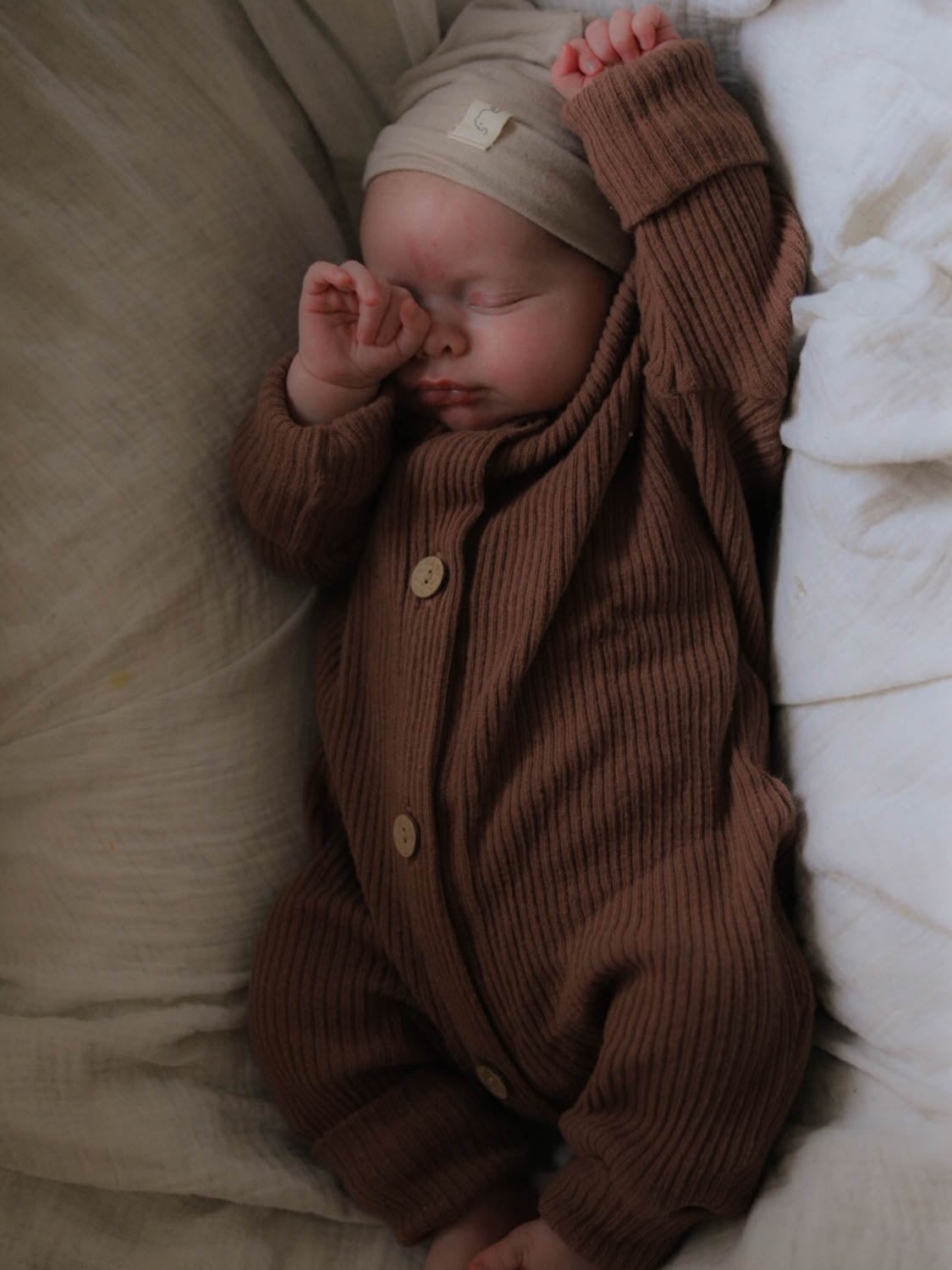 Mini Overall Rippstrick - Nougat: Ein Baby schläft in einem braunen, weichen Rippstrick-Overall aus recycelter Baumwolle, der Komfort und Wärme bietet. Erstausstattung Baby und Baby Geschenk zur Geburt.