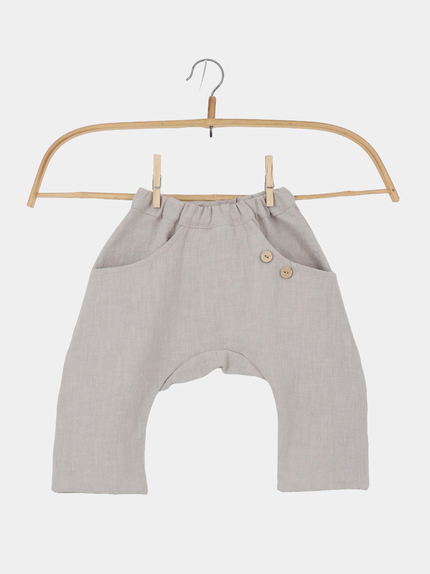 Sommerknicker Leinen - Sand: Leinenartige, hellgraue Baby-Haremshose mit elastischem Bund, tiefem Schnitt, vorderen aufgesetzten Taschen und zwei dekorativen Holzknöpfen, präsentiert an einem schlichten Holzkleiderbügel.
