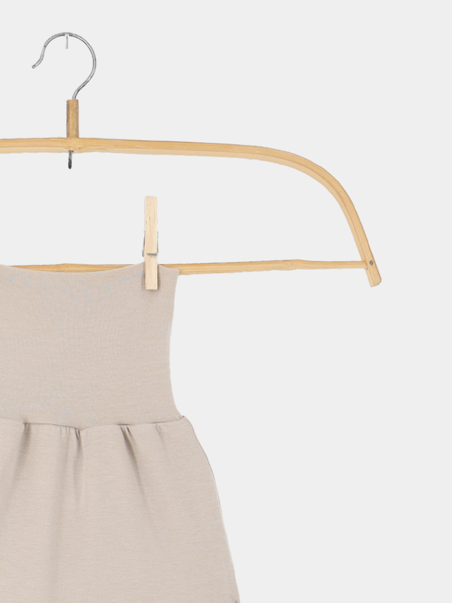 Baby Strampel- und Pucksack Kaschmir-Mix Sand hängt an einem natürlichen Holzbügel, mit einem kleinen Holzklammer gesichert, präsentiert die elegante, geraffte Struktur des leichten, beigen Kleidungsstücks.