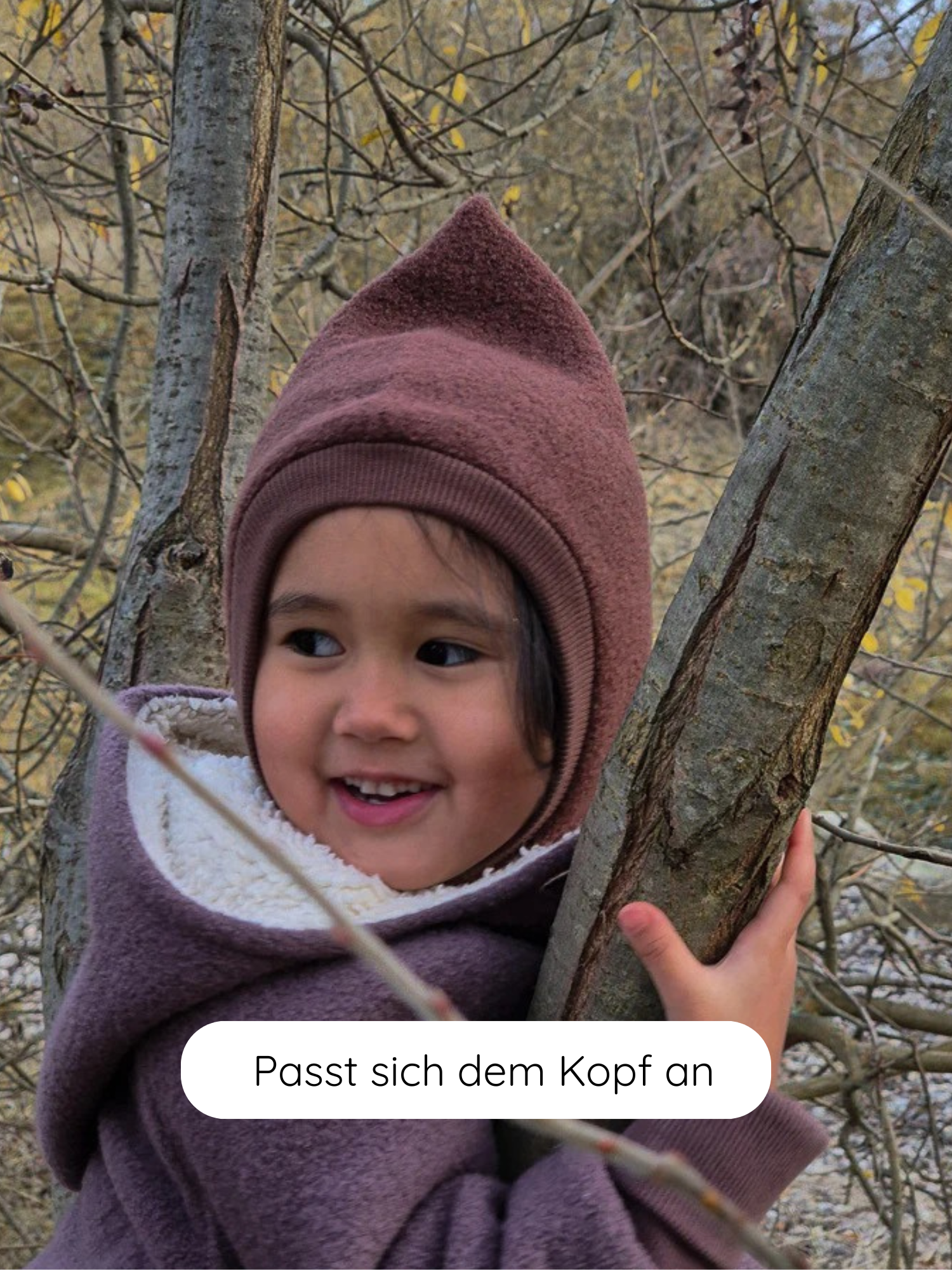 Ein Kind trägt eine Schlupfmütze Wollwalk - Schoko mit spitzem Kapuzen-Design, kuscheliger Fleece-Innenfutter und hält lächelnd einen Baumast fest. Der Text „Passt sich dem Kopf an.“ ist sichtbar.