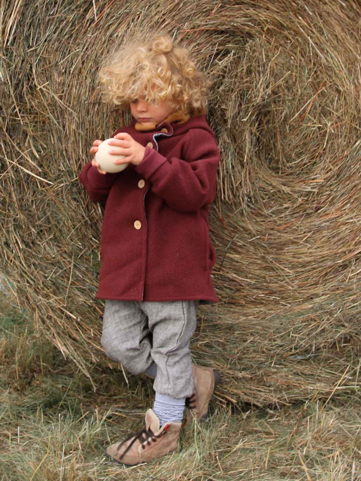 Kind mit Wichteljacke Wollwalk - Ziegel hält weißen Ball, steht vor Strohballen. Jacke aus Wollwalk, gefüttert mit Bio-Jersey, ideal für Herbst bis Frühling, bietet Wärme und Bewegungsfreiheit.