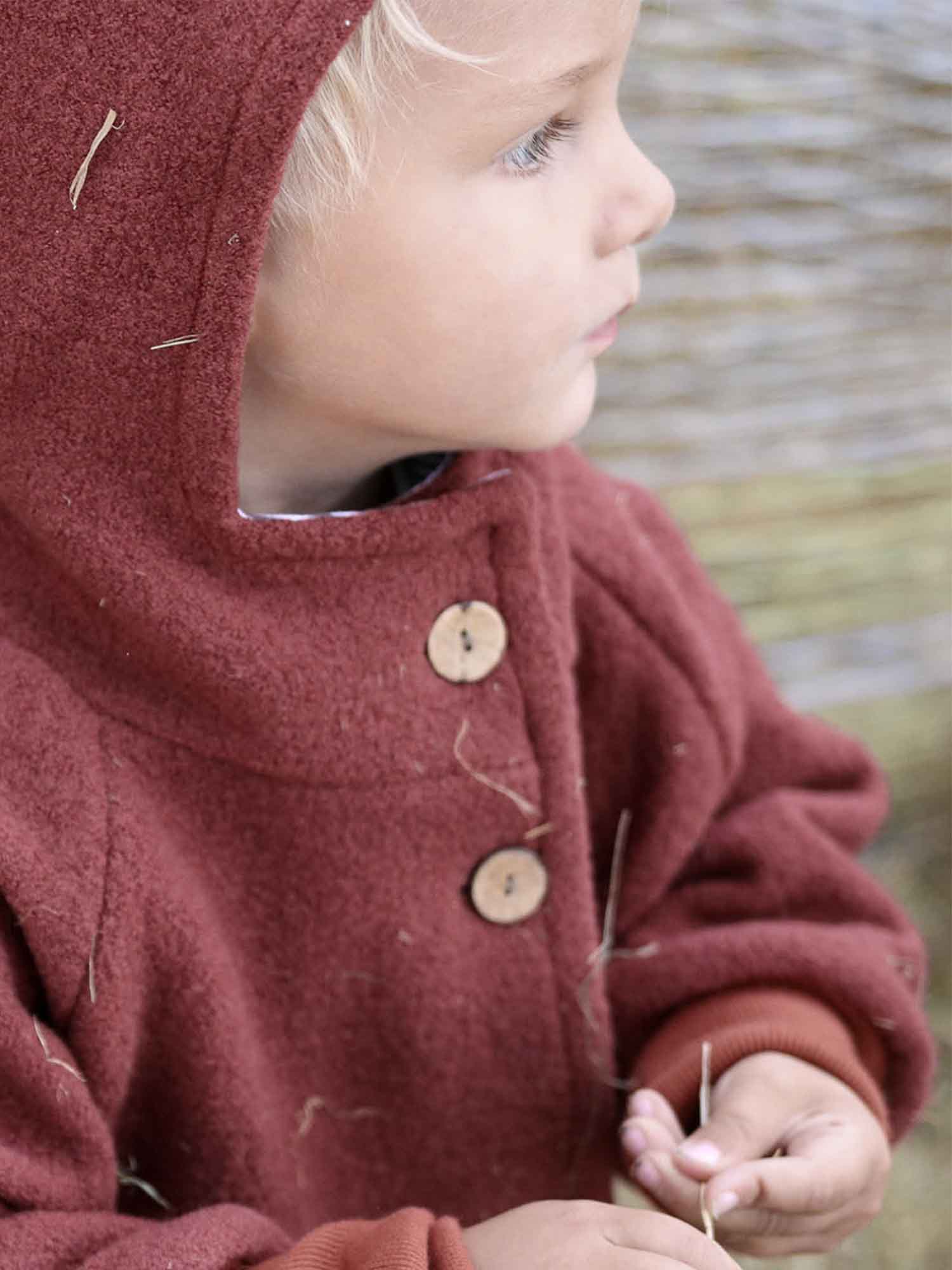 Kleinkind im Profil mit rust-roter Wichteljacke Wollwalk - Ziegel, Kapuze, zwei runde Holzknöpfe, hält Stroh. Jacke aus Wollwalk, ideal für kalte Jahreszeiten, bietet Bewegungsfreiheit und Komfort.