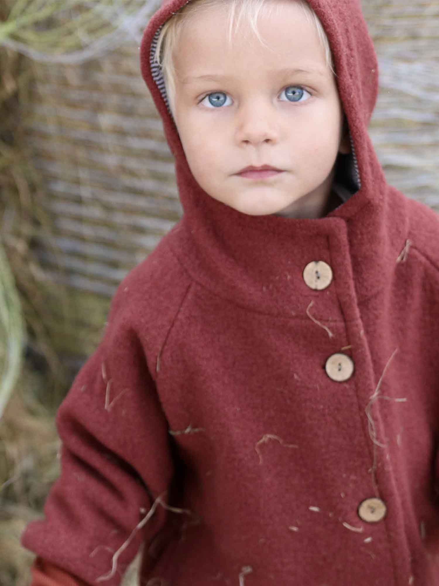 Kleinkind in Wichteljacke Wollwalk - Ziegel mit Kapuze und Holzknöpfen, draußen stehend. Die Jacke zeigt Heuflecken und bietet tragefreundliche Wärme durch Bio-Baumwollfutter, ideal für kühle Jahreszeiten.