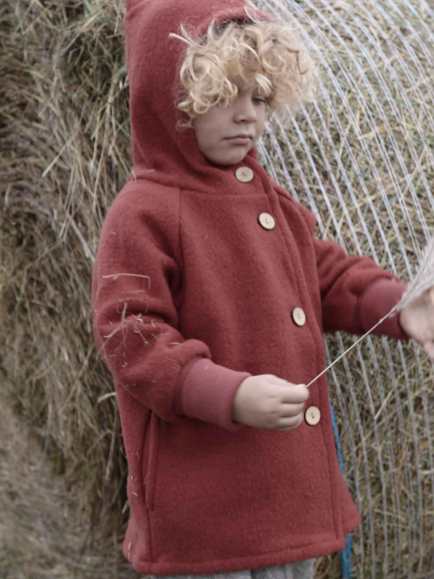 Kleinkind mit lockigem Haar trägt die Wichteljacke Wollwalk - Ziegel und steht neben einem Heuballen, hält einen Strohhalm in der Hand, ideal für kalte Jahreszeiten.