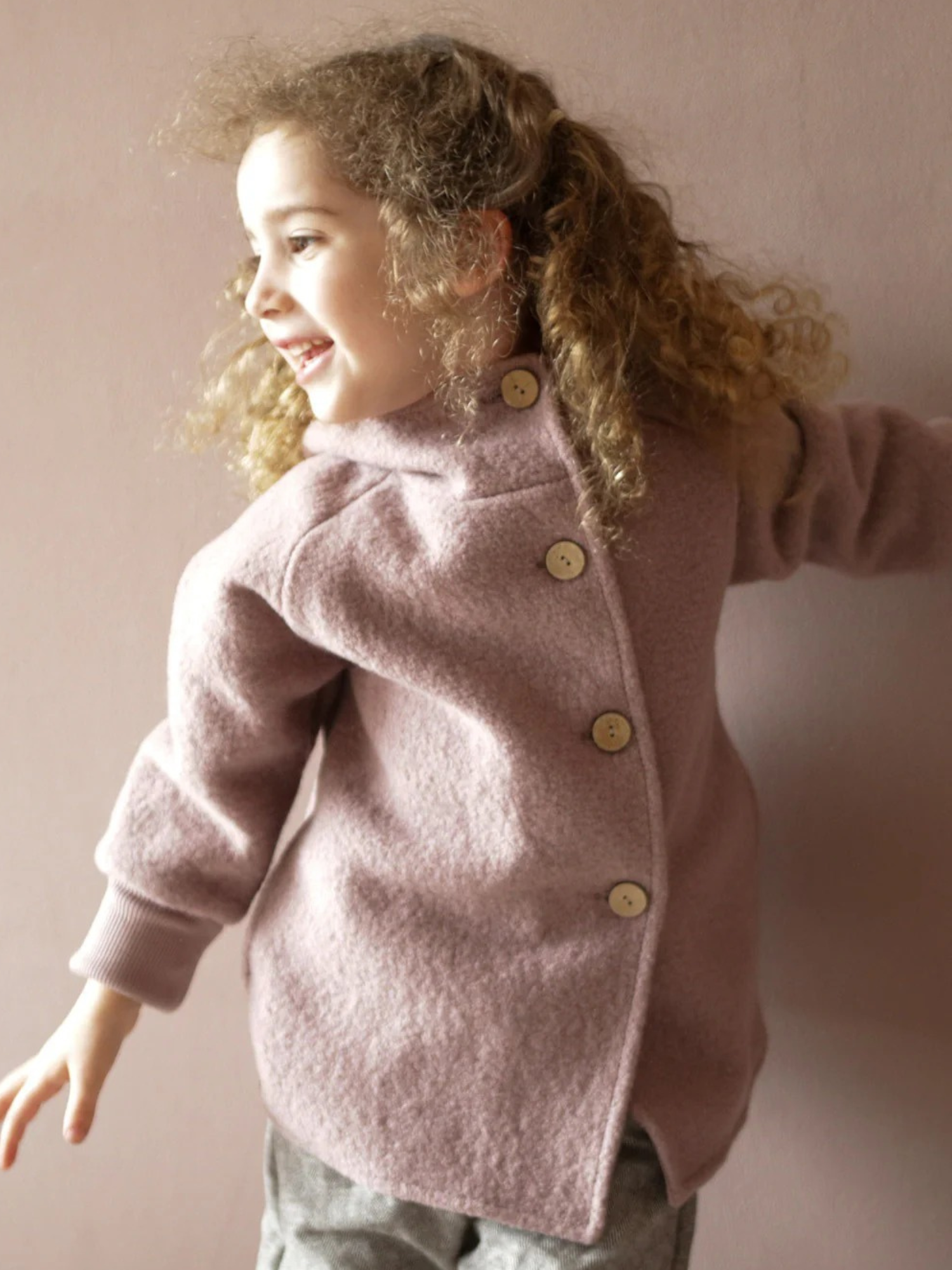Kind trägt die Wichteljacke Wollwalk - Puder mit vier großen Holzknöpfen am Rücken. Die staubrosafarbene Jacke hat einen mitwachsenden Schnitt und ist aus weichem Wollwalk gefertigt.