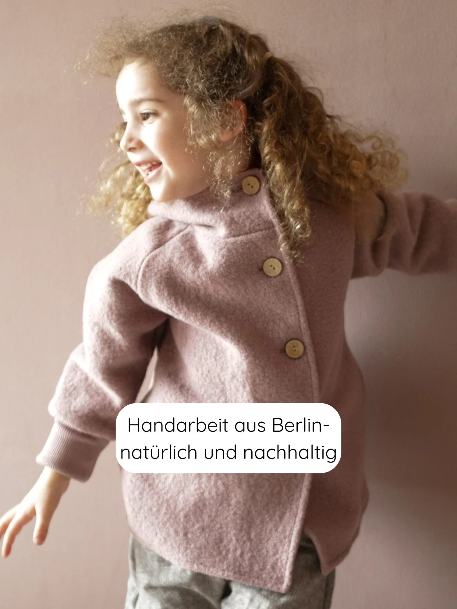 Ein lächelndes Kind trägt die Wichteljacke Wollwalk - Puder mit Holzknöpfen hinten, kombiniert mit grauen Hosen. Die Jacke ist handgefertigt, nachhaltig und aus Berlin.