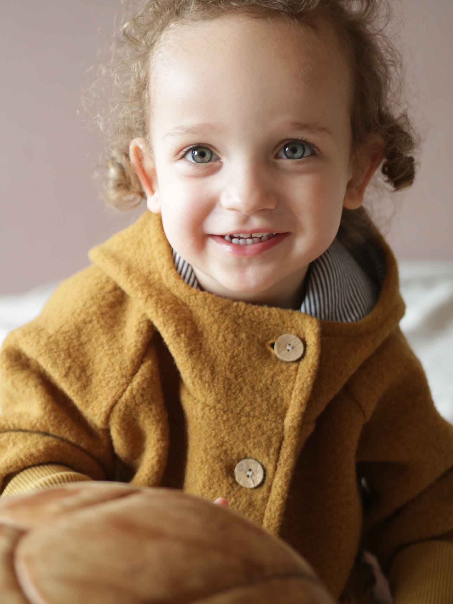 Ein lächelndes Kleinkind trägt die Wichteljacke Wollwalk - Honig mit Holzknöpfen, hält eine braune Lederkugel. Die Jacke ist gefüttert und ideal für Herbst, Winter und Frühling.