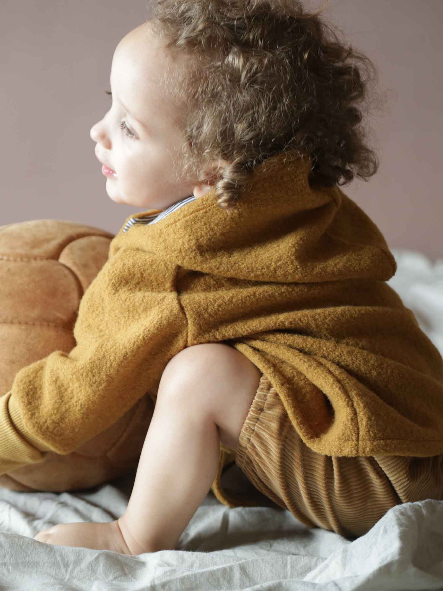 Kleinkind trägt die Wichteljacke Wollwalk - Honig. Die warme, kapuzenförmige Jacke mit Bio-Jersey-Futter bietet Bewegungsfreiheit, ideal für Herbst. Sie ist aus hochwertigem Wollwalk, perfekt für kalte Tage.