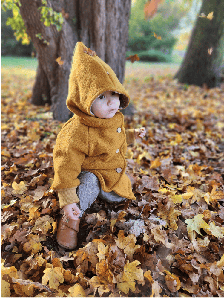 Kleinkind in Wichteljacke Wollwalk - Honig, gebückt im Herbstlaub. Die handgemachte Jacke mit spitzem Kapuze bietet Bewegungsfreiheit und Komfort, ideal für kühlere Tage.