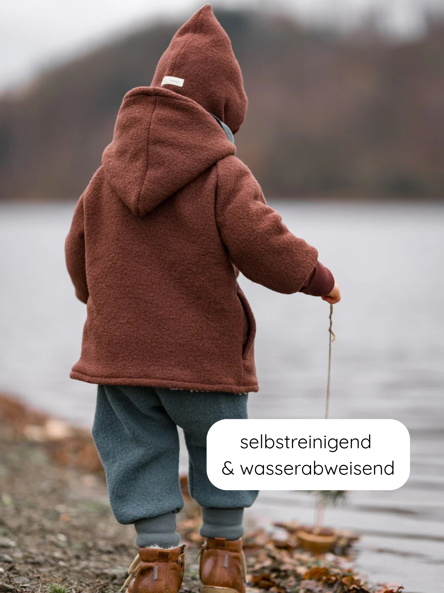 Ein Kind in einer Wichteljacke Wollwalk - Schoko steht am Wasser, hält einen Stock. Der Mantel ist selbstreinigend, wasserabweisend und ideal für Herbst, Winter und Frühling.