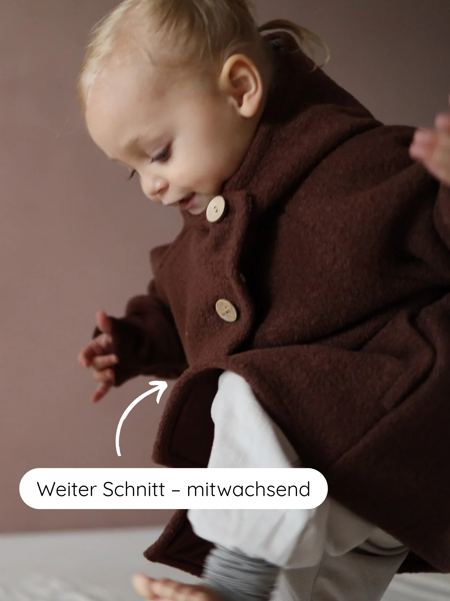 Ein Kleinkind trägt die Wichteljacke Wollwalk - Schoko mit weitem Schnitt, der mitwächst. Ein Pfeil zeigt auf den Saum mit dem Label „Weiter Schnitt – mitwachsend“.