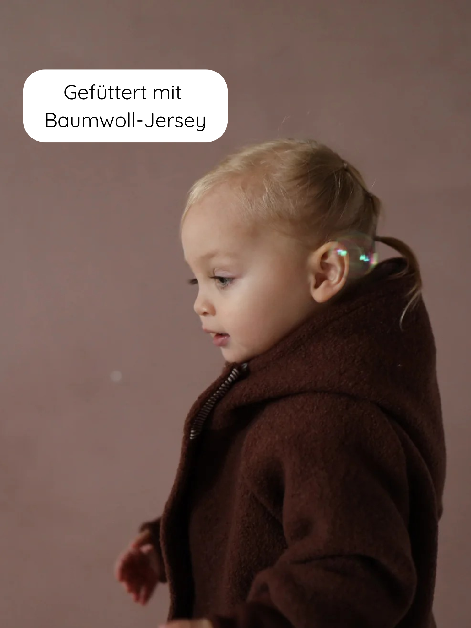 Ein Kleinkind trägt die Wichteljacke Wollwalk - Schoko aus Wollwalk mit Kapuze, die mit Bio-Baumwolljersey gefüttert ist. Eine schwebende Seifenblase ist neben dem Ohr sichtbar.