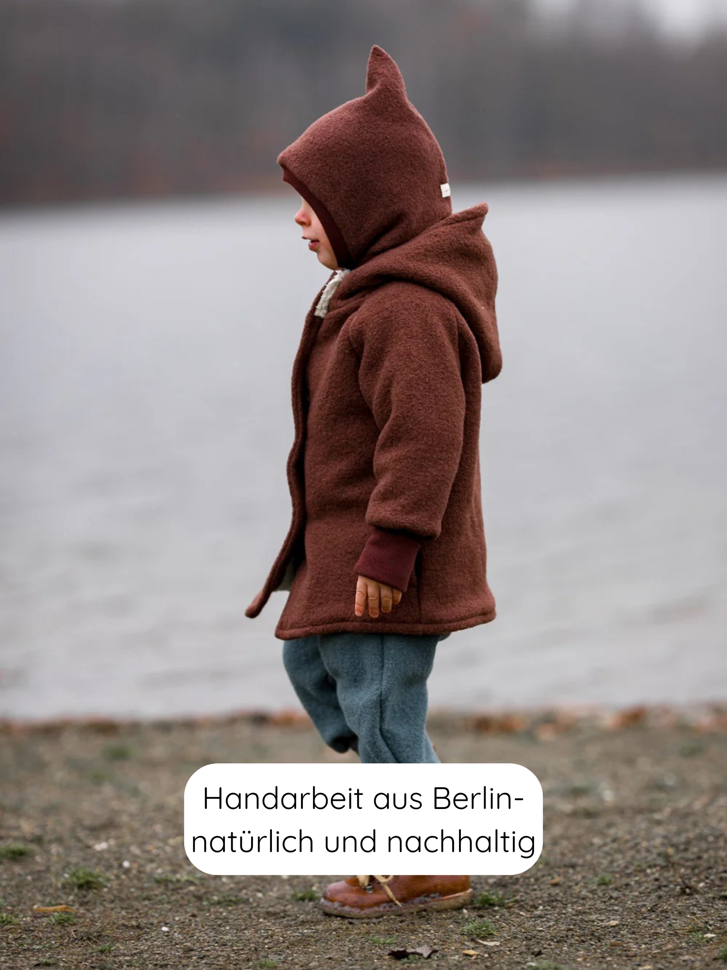 Ein Kleinkind steht am Ufer, trägt eine Wichteljacke Wollwalk - Schoko mit spitzem Kapuzenabschluss, ideal für Herbst bis Frühling, handgefertigt in Berlin, warm und nachhaltig.