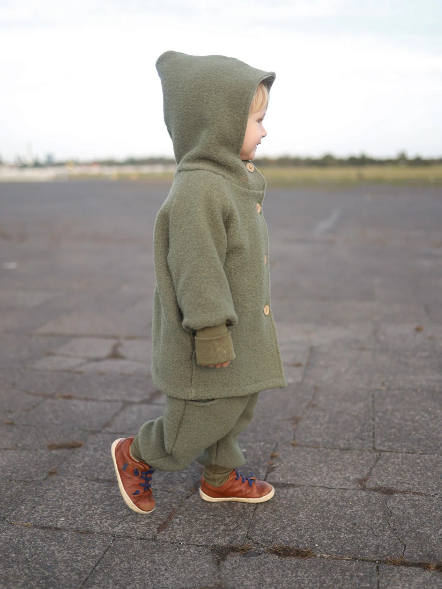Ein Kleinkind trägt die Wichteljacke Wollwalk - Pistazie mit einem Kapuzenmantel und Holzknöpfen, kombiniert mit passenden Hosen und braunen Sneakers mit blauen Schnürsenkeln, auf einem weiten, gepflasterten Boden.