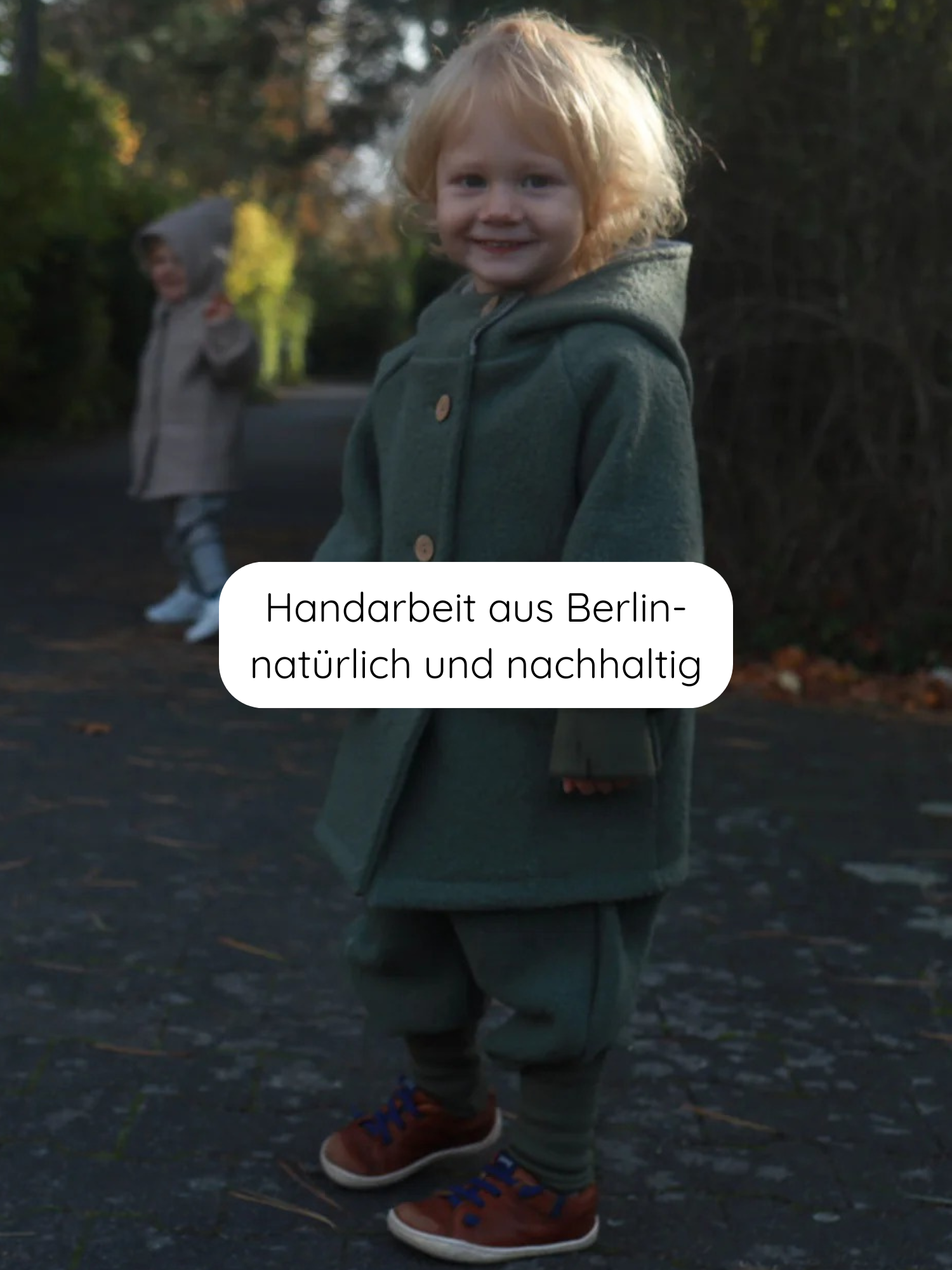 Zwei Kinder im Freien, im Fokus ein lächelnder Kleinkind. Es trägt die Wichteljacke Wollwalk - Pistazie, eine grüne, handgefertigte, nachhaltige Jacke mit Kapuze. Text: “Handarbeit aus Berlin – natürlich und nachhaltig”.
