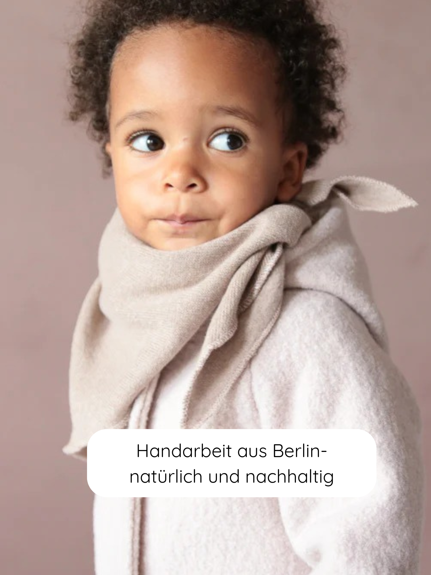 Kleinkind mit lockigem Haar trägt die Wichteljacke Wollwalk - Sand mit einem weichen, beigefarbenen Schal. Im Vordergrund ein Etikett: Handarbeit aus Berlin – natürlich und nachhaltig.