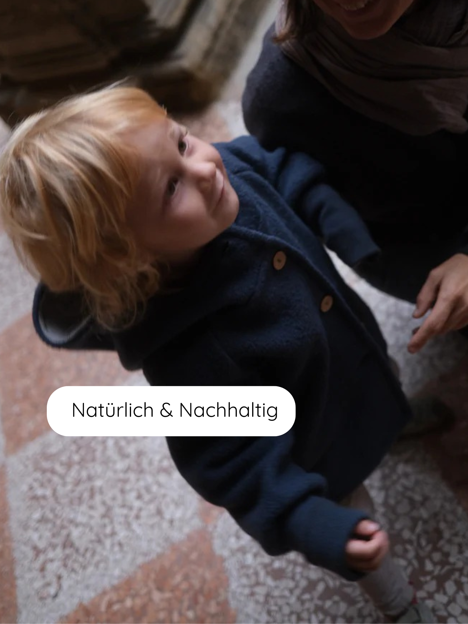 Kind in dunkelblauer Wichteljacke Wollwalk - Marine, auf Steinboden stehend. Ein teilweise sichtbarer Erwachsener greift nach dem Kind. Auf dem Bild steht „Natürlich & Nachhaltig“.